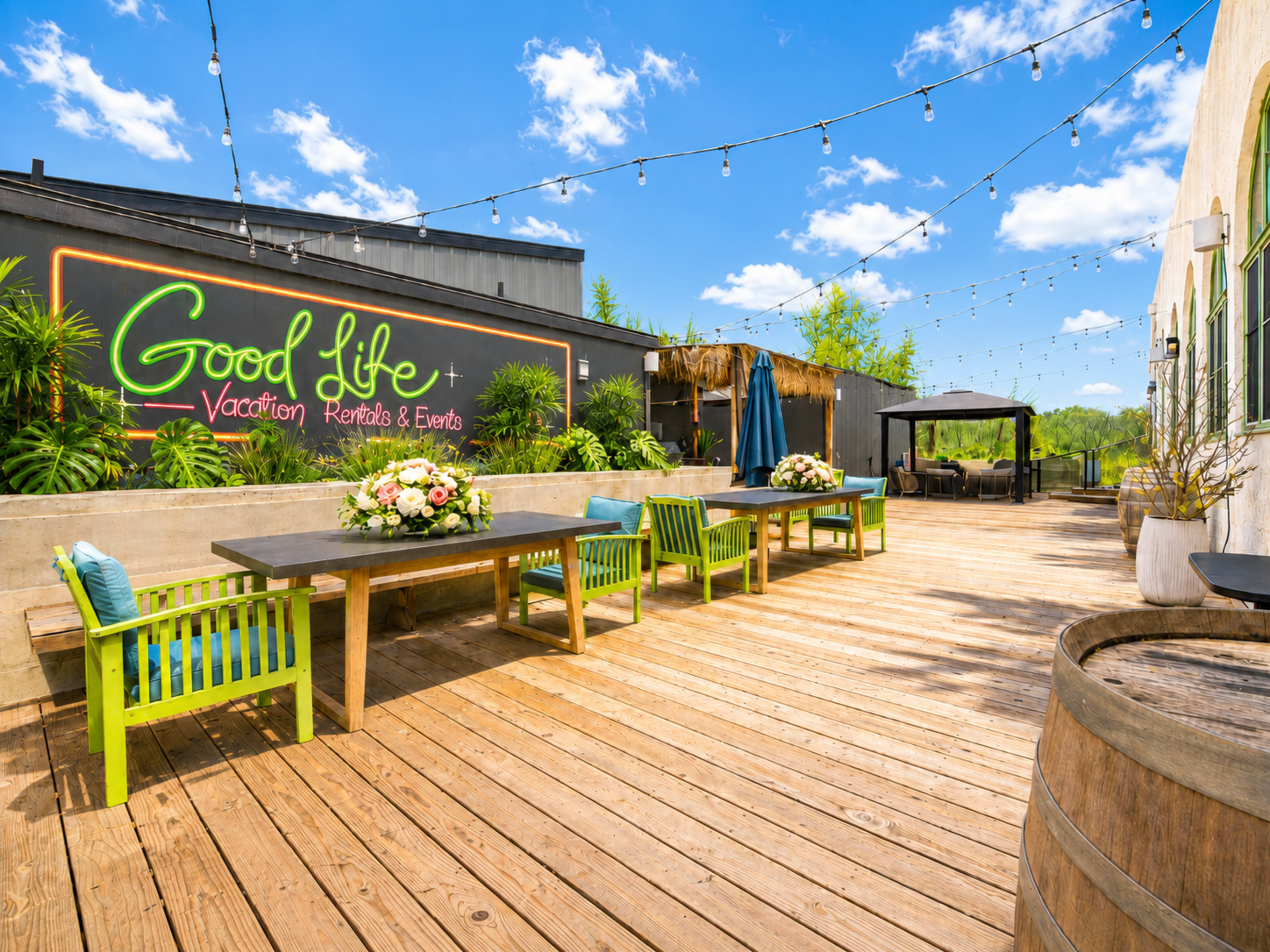 The image shows a spacious outdoor area with wooden flooring, green and blue chairs, floral table centerpieces, and string lights overhead, featuring a colorful mural that reads "Good Life - Vacation Rentals & Events."