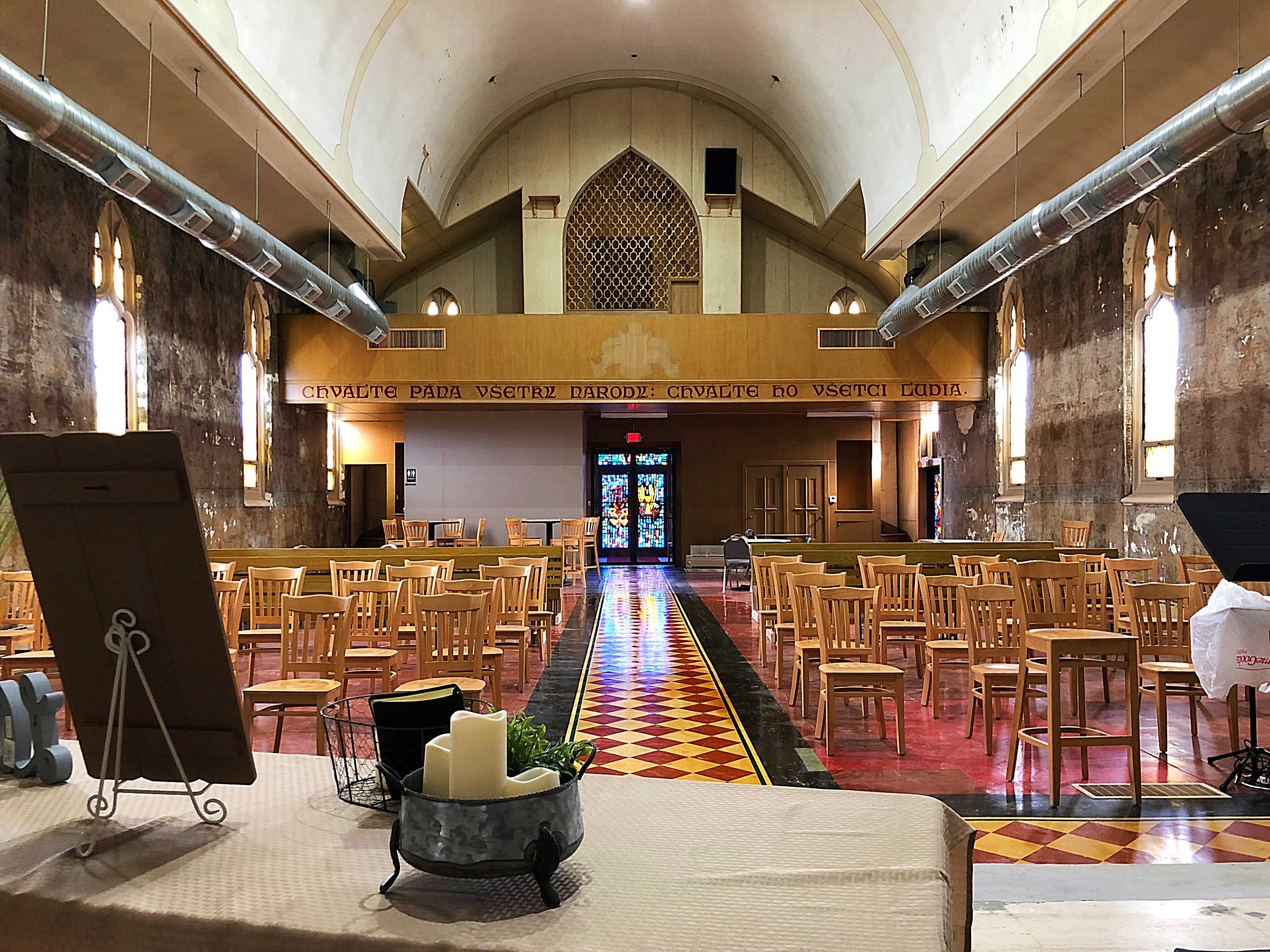 The interior of a large, empty church with wooden chairs arranged in rows and a colorful stained glass window at the far end.