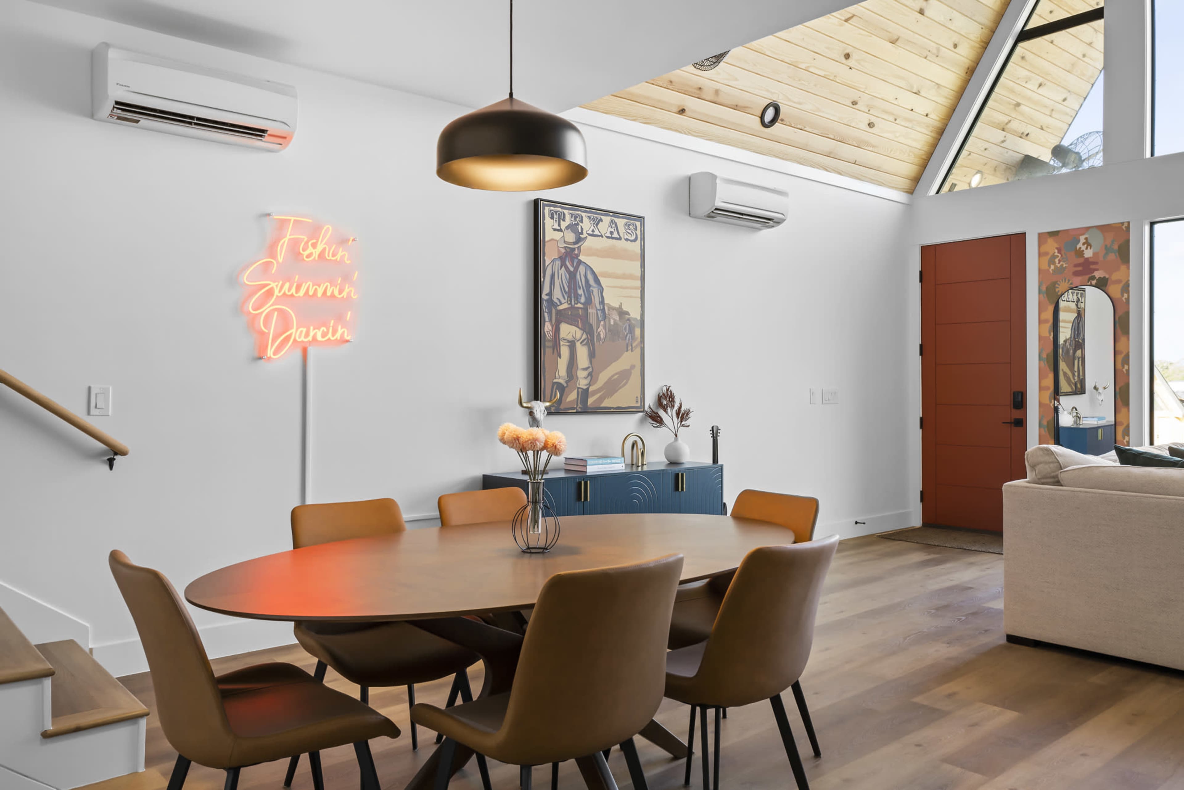 A dining area features a round table with eight chairs, a neon sign, and a large artwork on the wall, illuminated by natural light from a skylight.