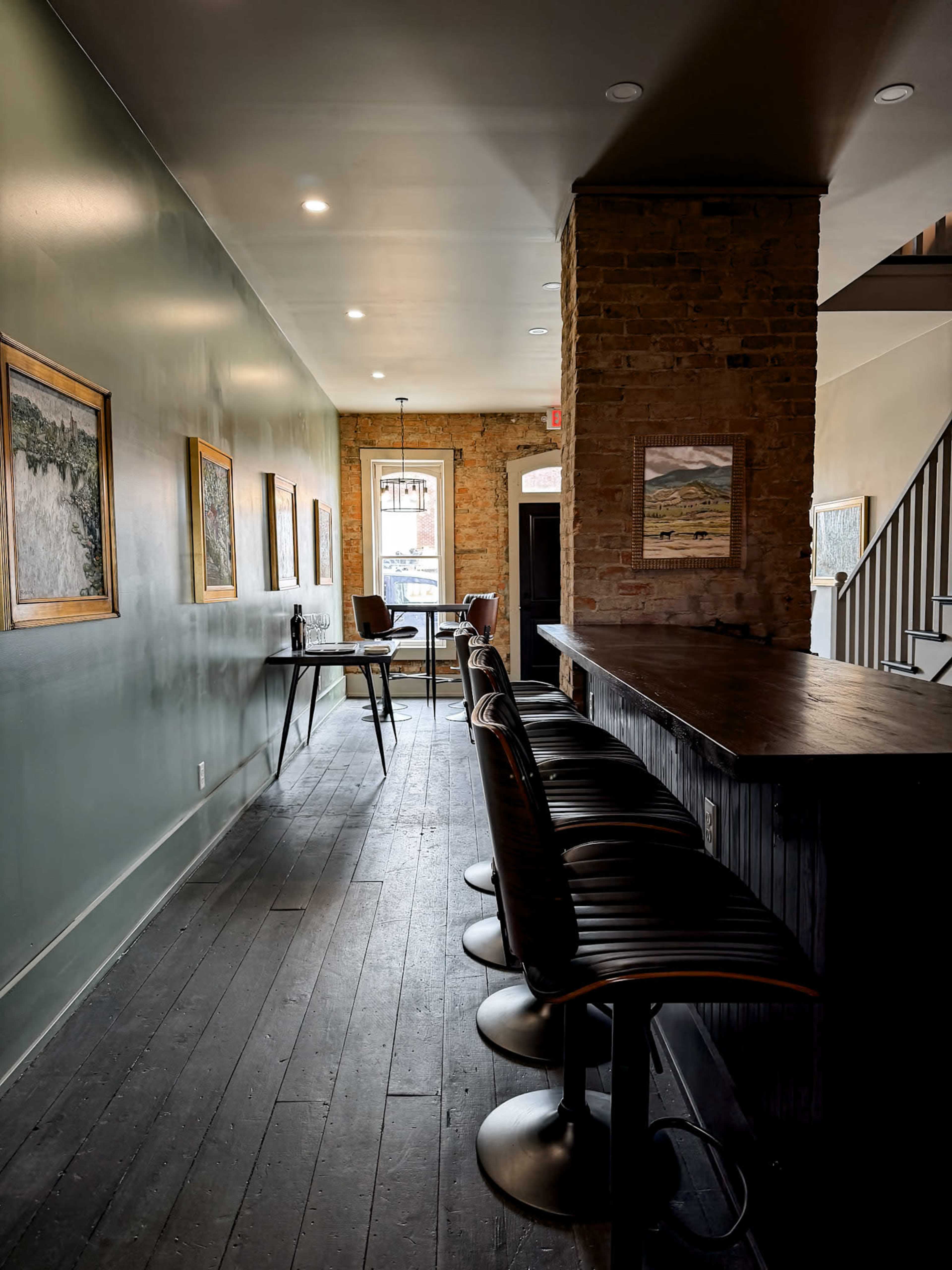 The image shows a narrow hallway with a bar lined with stools on one side and framed artwork along the walls, leading to a window at the far end.