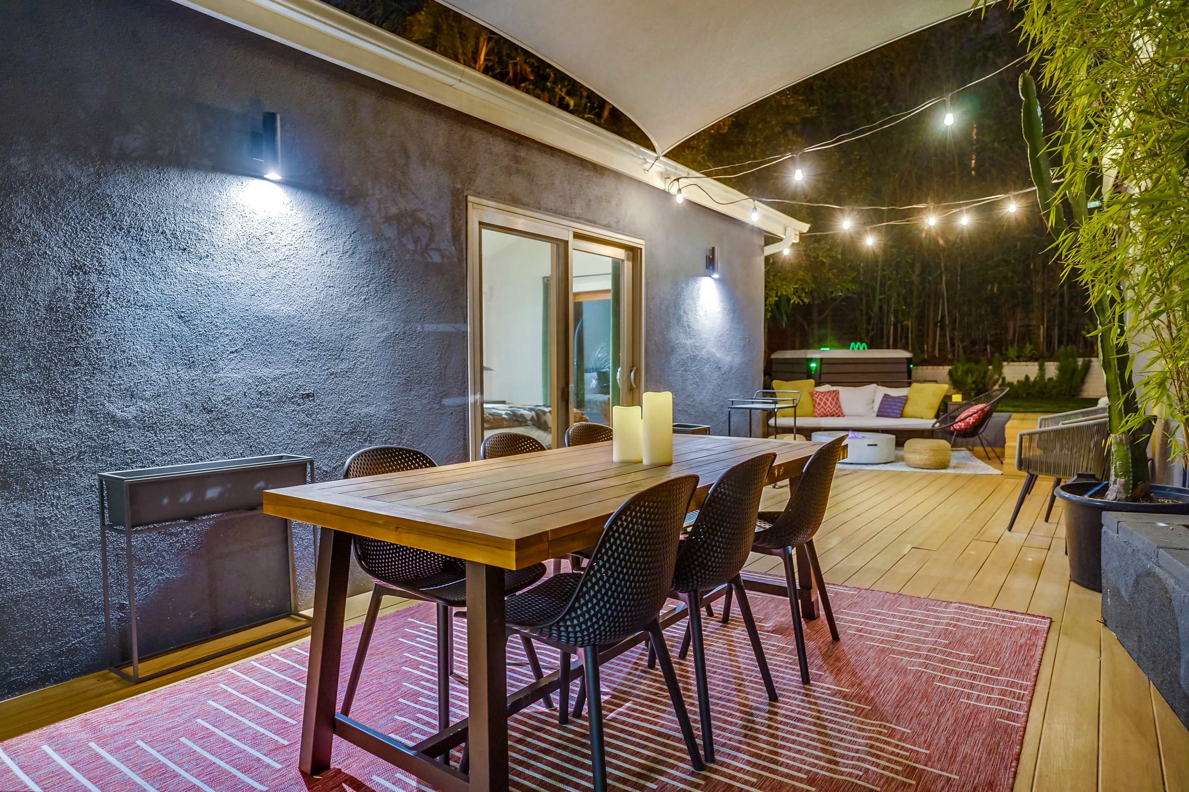 The image shows a modern outdoor dining area featuring a wooden table surrounded by black chairs, with soft lighting and a cozy atmosphere.