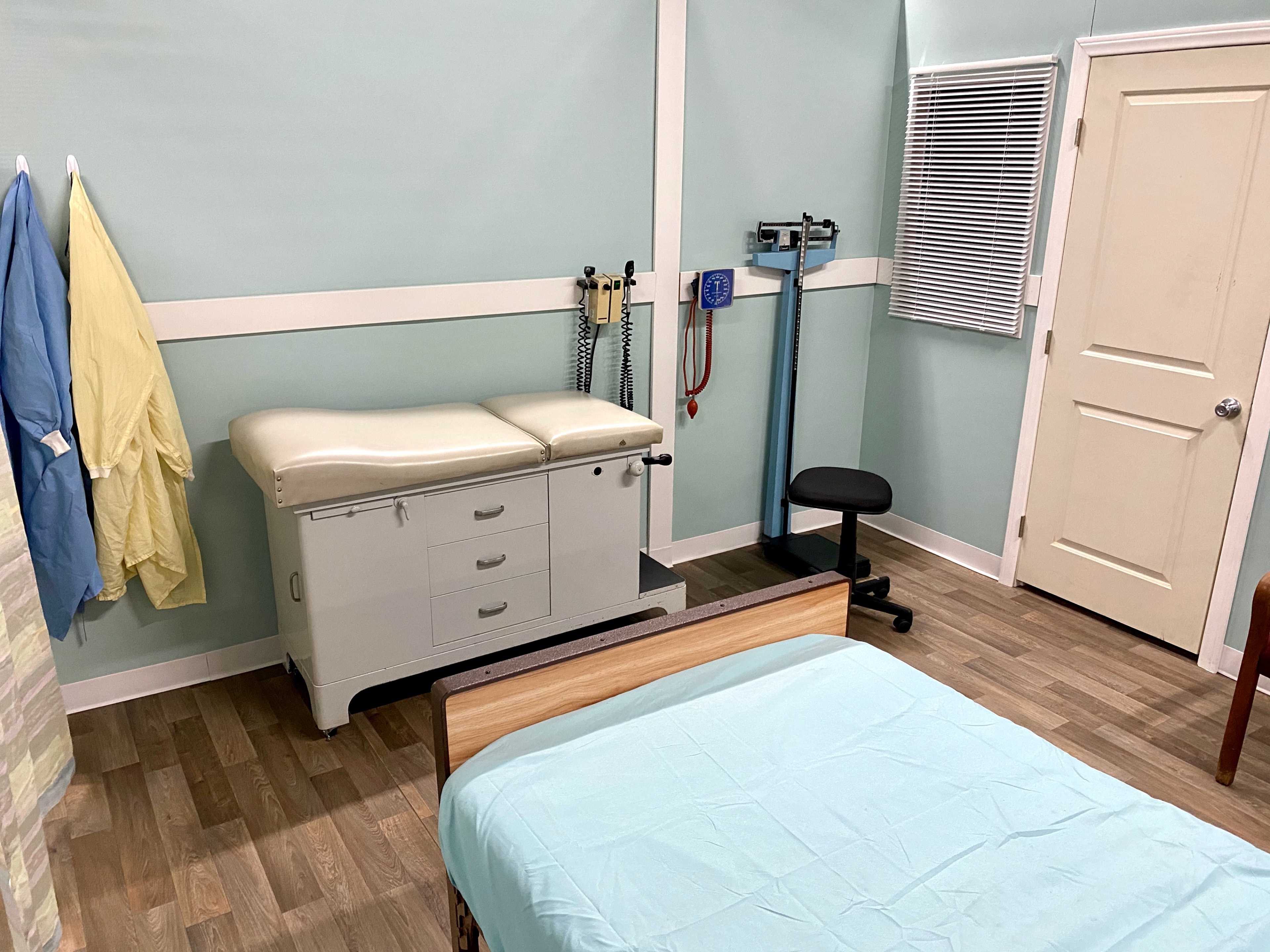 A medical examination room with a bed, examination table, medical equipment, and a chair.