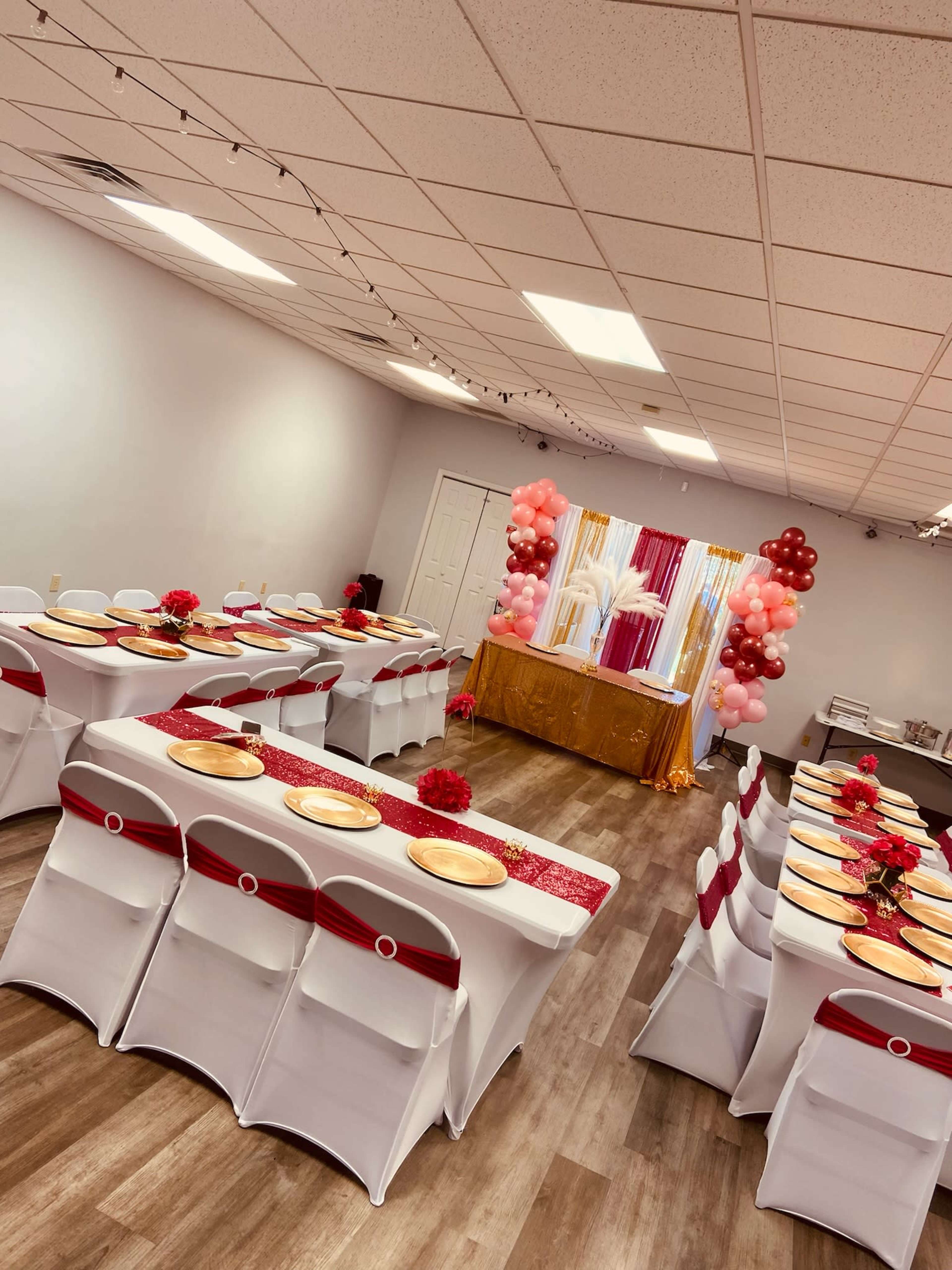 A decorated event space with rows of tables set with white tablecloths and red accents, balloons, and a backdrop of gold and pink decorations.