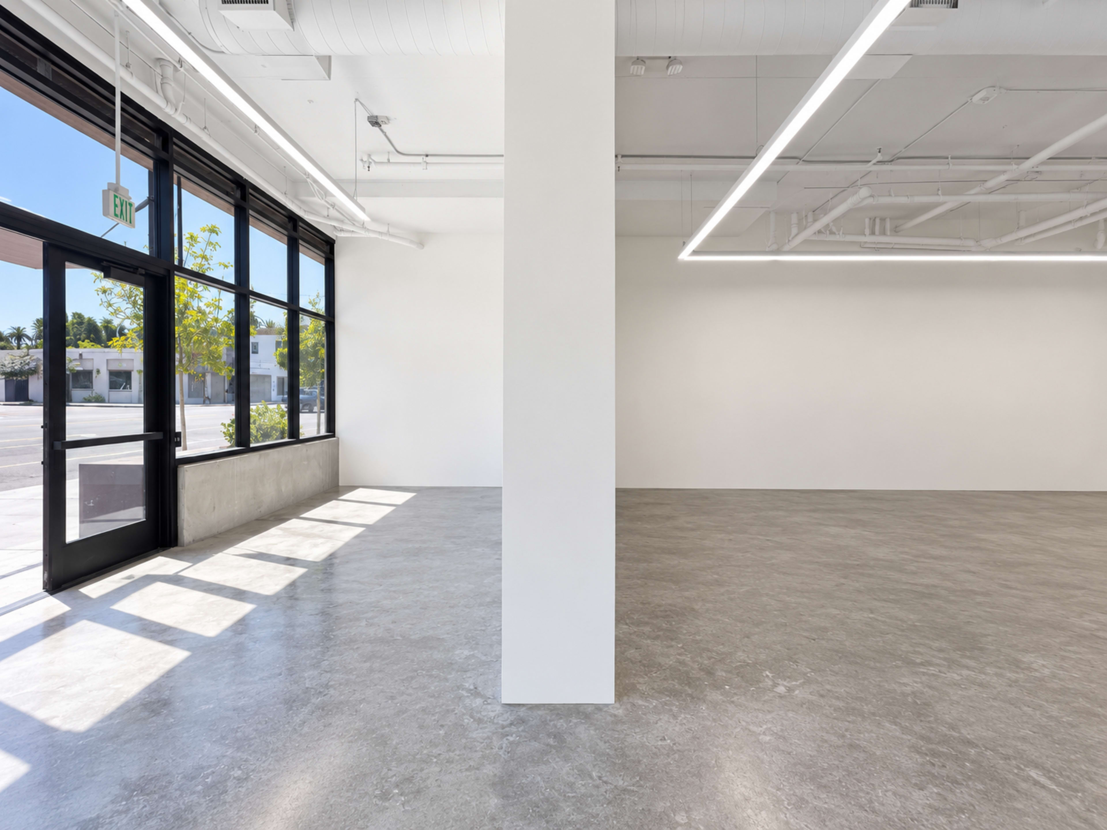 The image shows a spacious, modern interior with large windows, a concrete floor, and a central white column dividing the room.