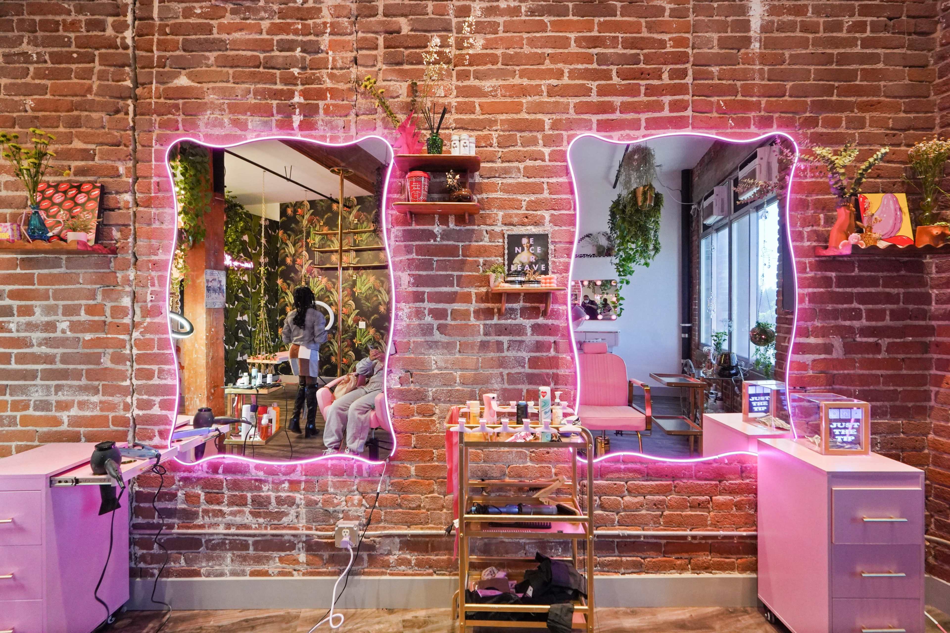 A salon interior with two illuminated mirrors framed in pink, set against a brick wall, alongside a styling station and plants.