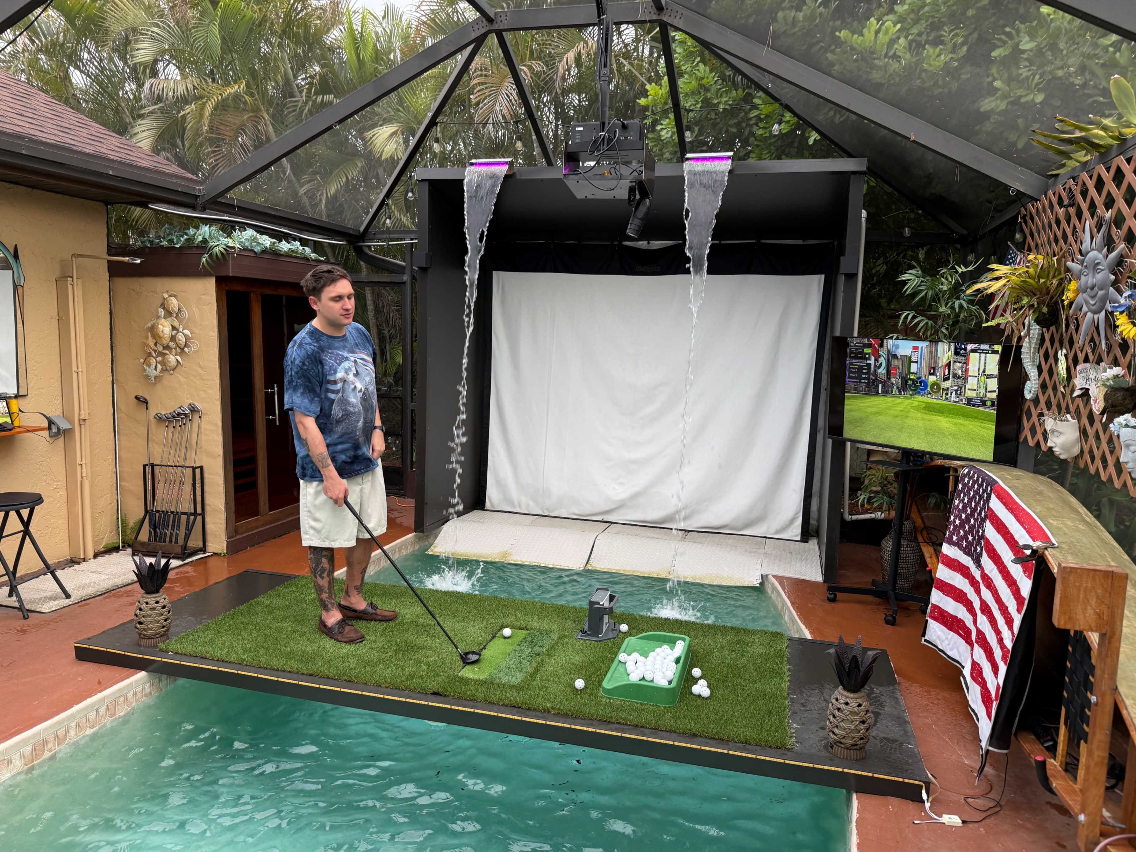 A man stands on a putting green in a covered outdoor area with a golf simulator screen and artificial turf, surrounded by greenery and pool water.