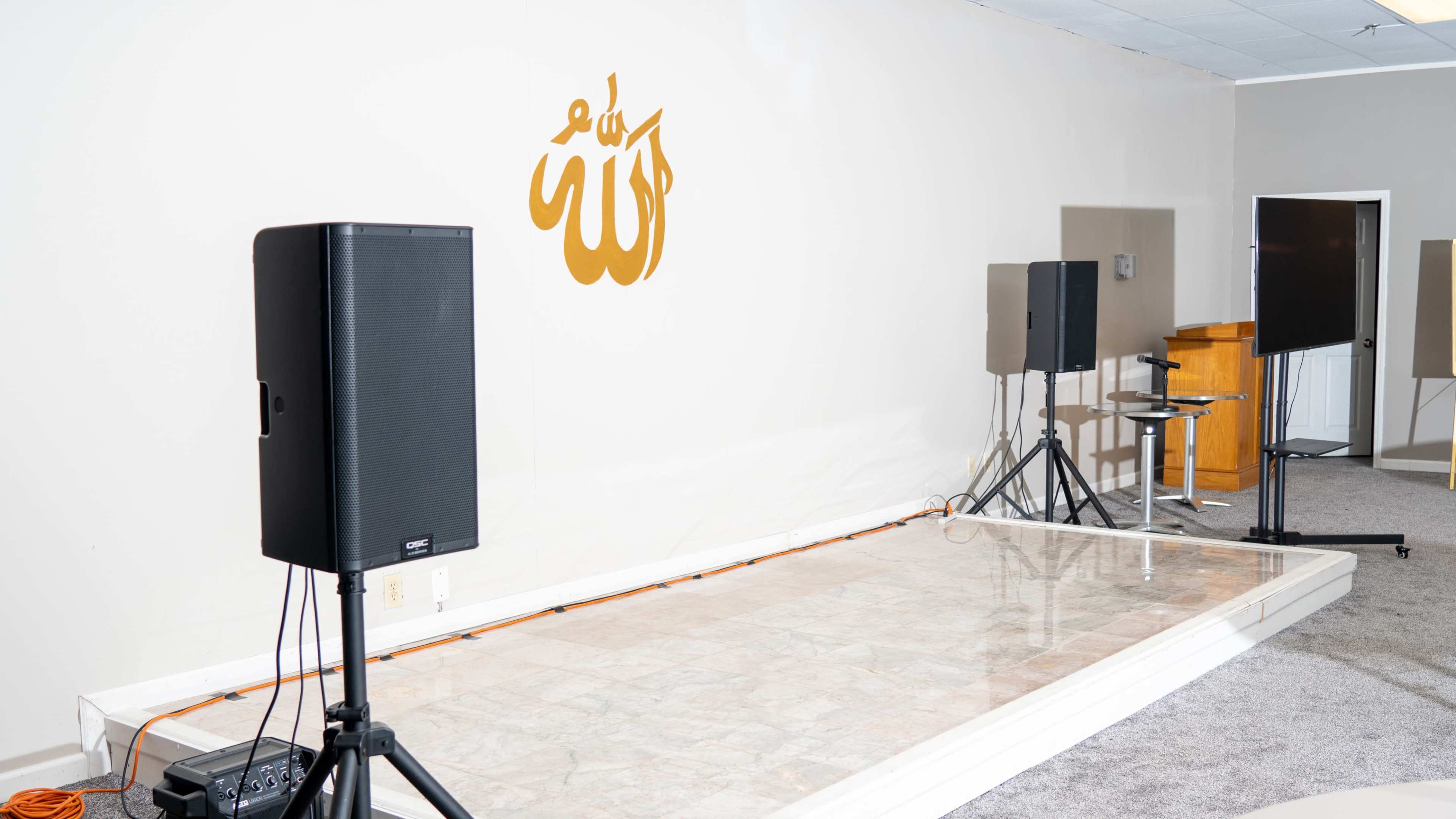 A small stage area with two speakers and a wooden podium against a plain wall featuring a decorative golden text.
