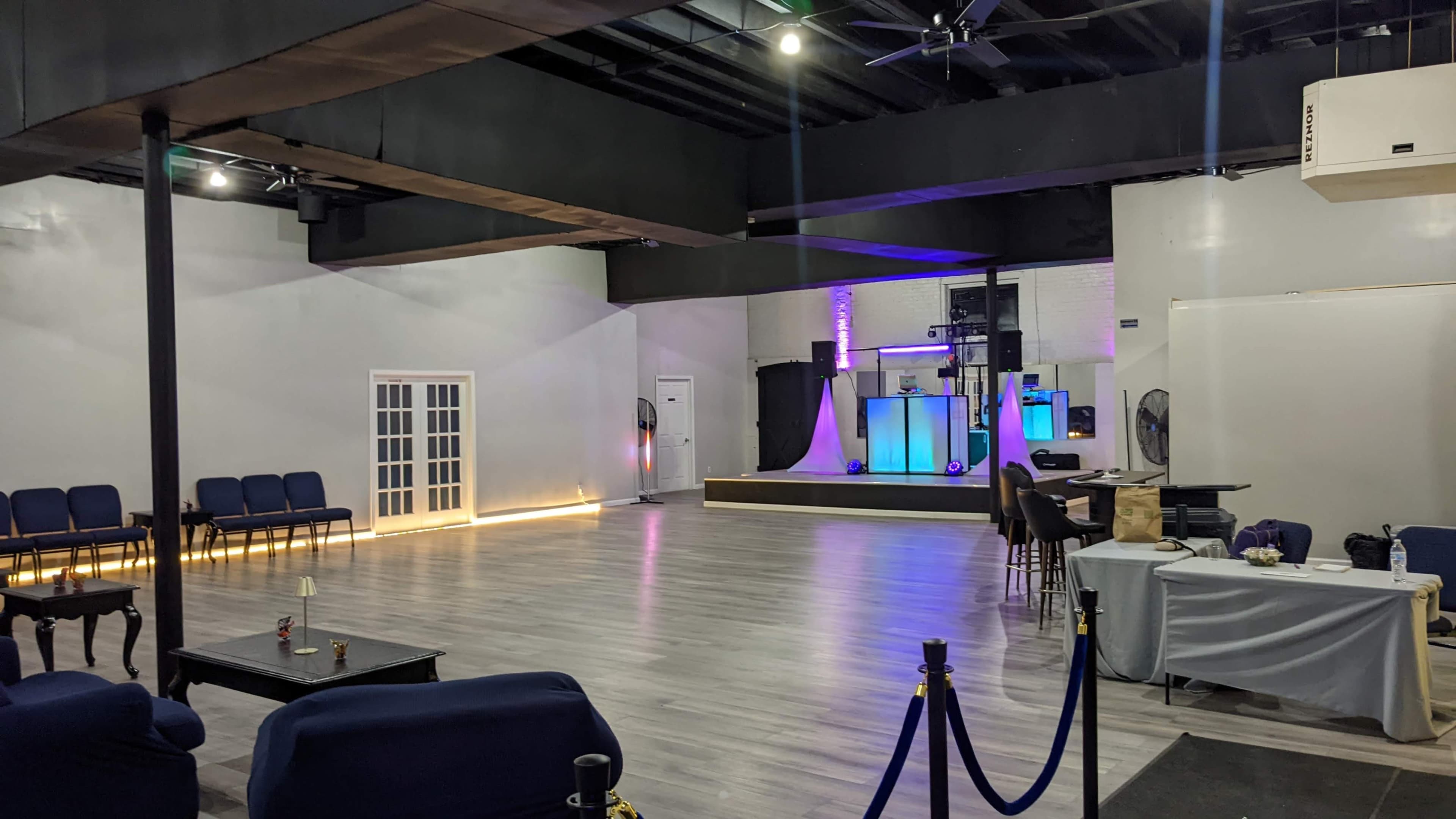A spacious event venue with seating arranged against the walls, a small stage illuminated at the front, and a bar area on the right.