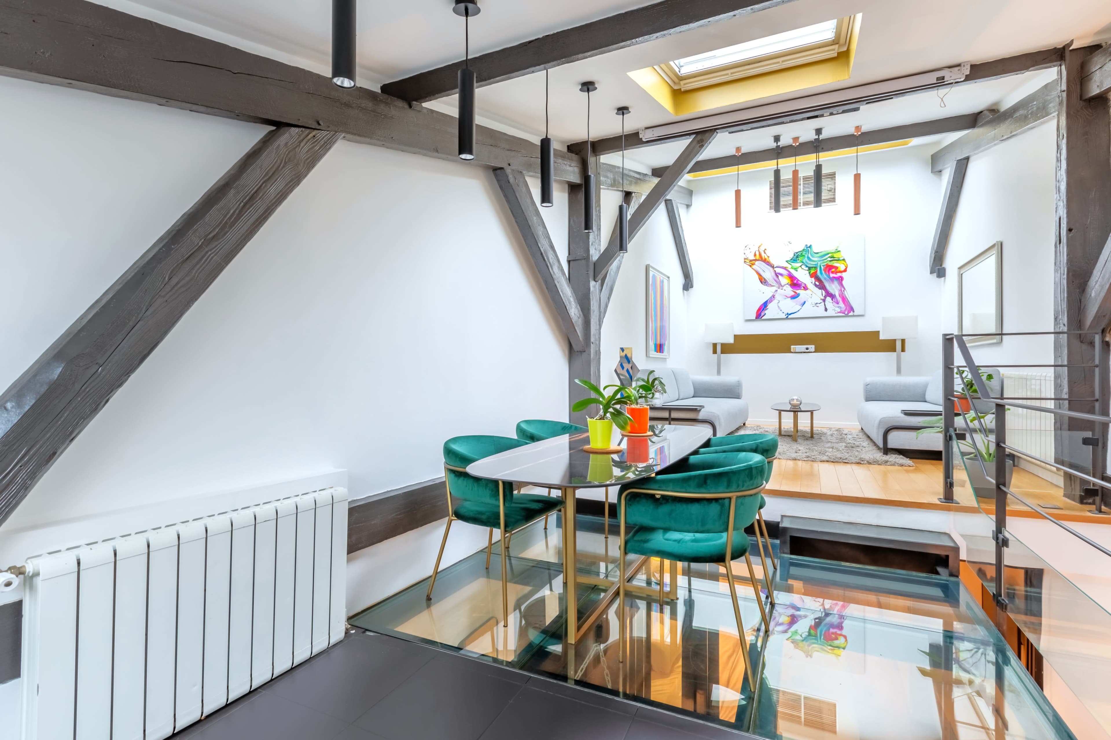 A modern interior features a dining area with a glass top table and green chairs, overlooking a living space with gray sofas and a colorful art piece on the wall.