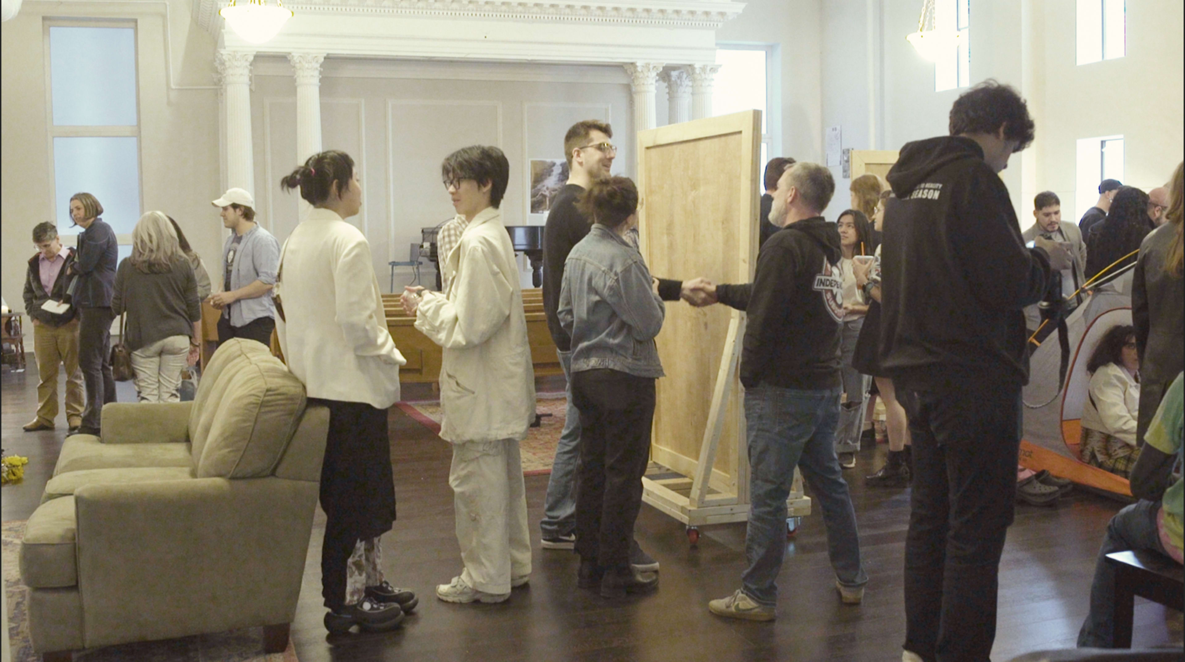 A group of people socialize and interact in a spacious room with light-colored walls and wooden floors.