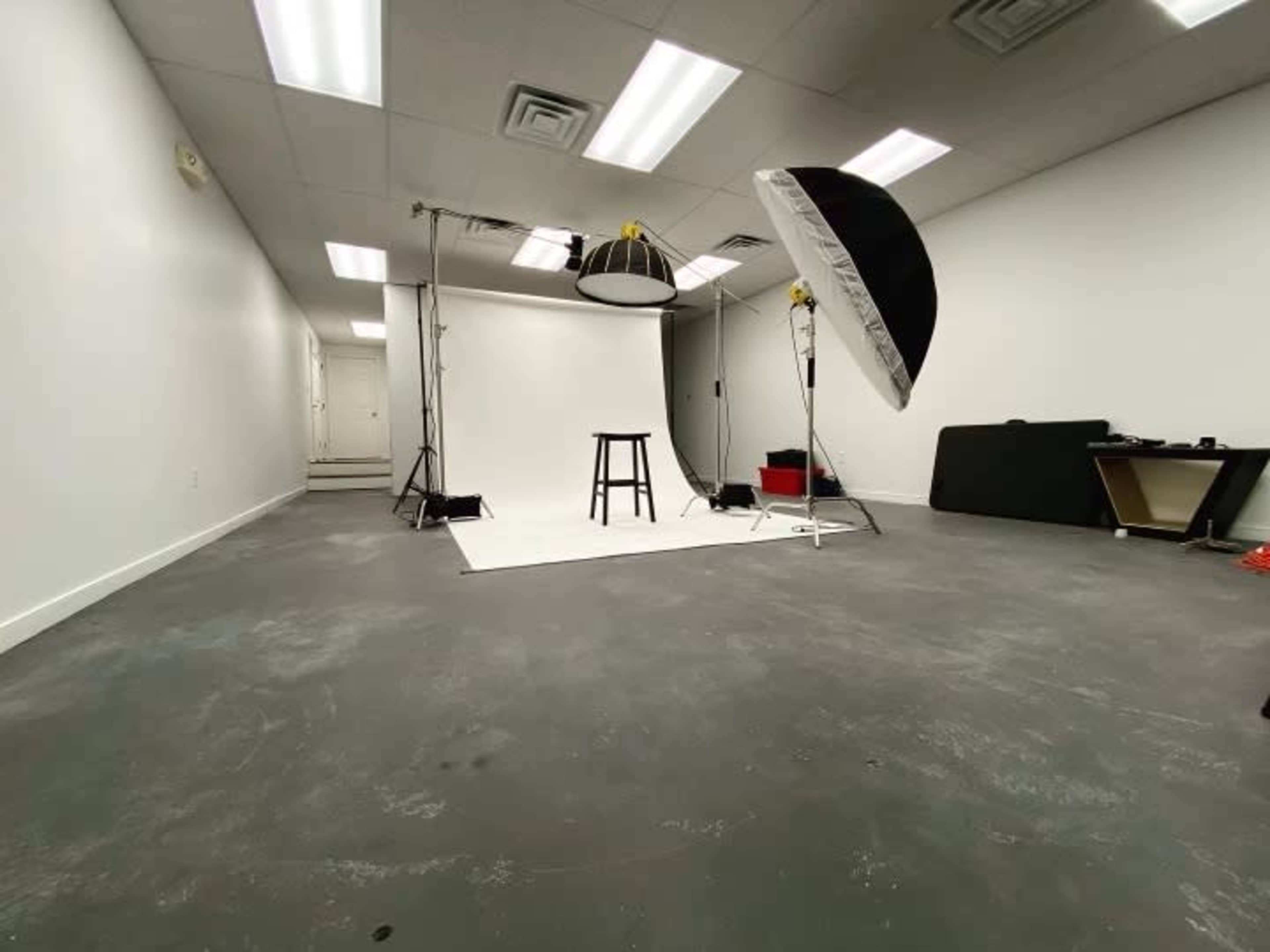 A photography studio with a white backdrop, lighting equipment, and a stool set up in an empty room.