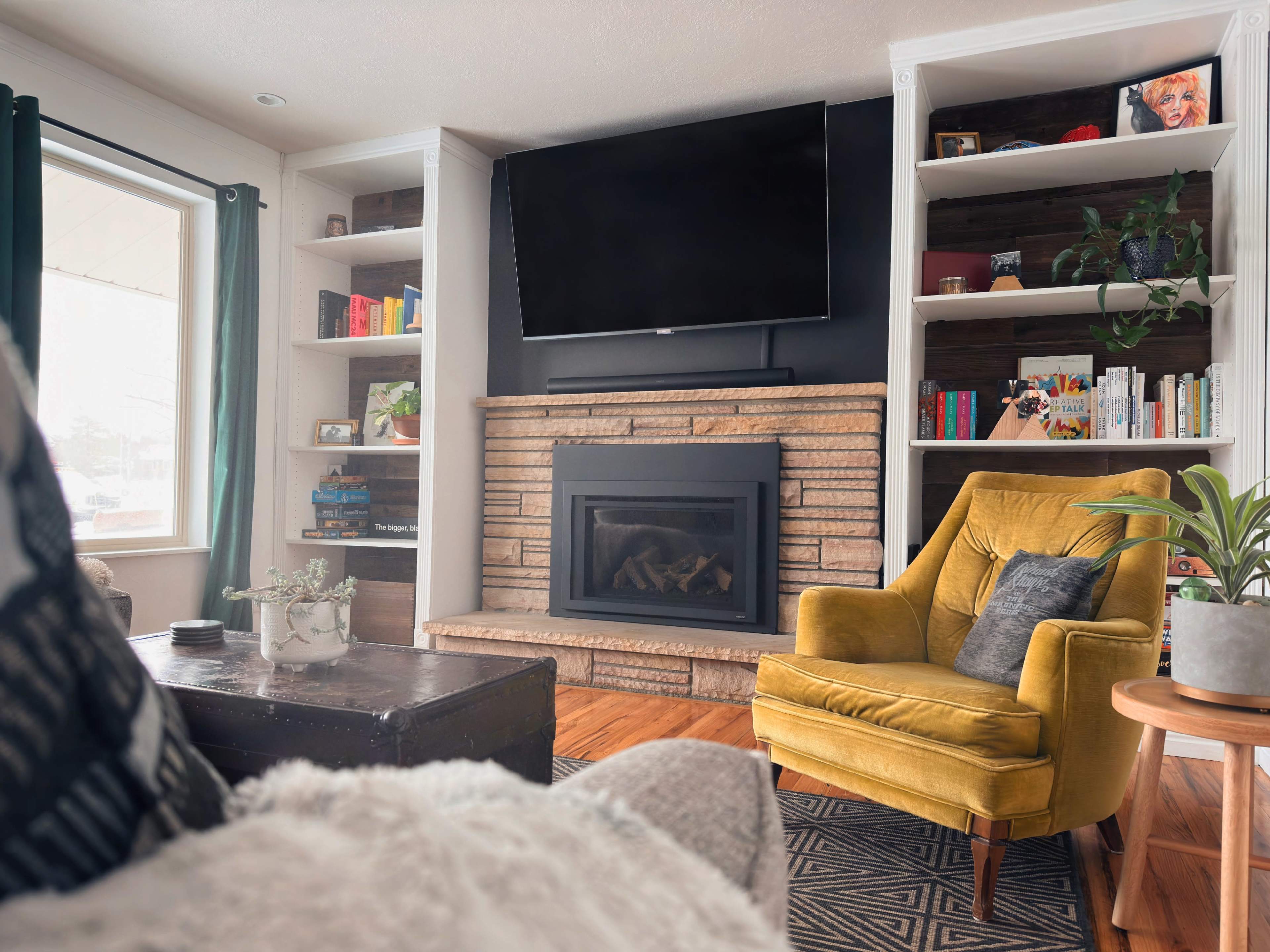 A cozy living room featuring a stone fireplace, a mounted TV, a yellow armchair, and built-in bookshelves filled with books and decorative items.