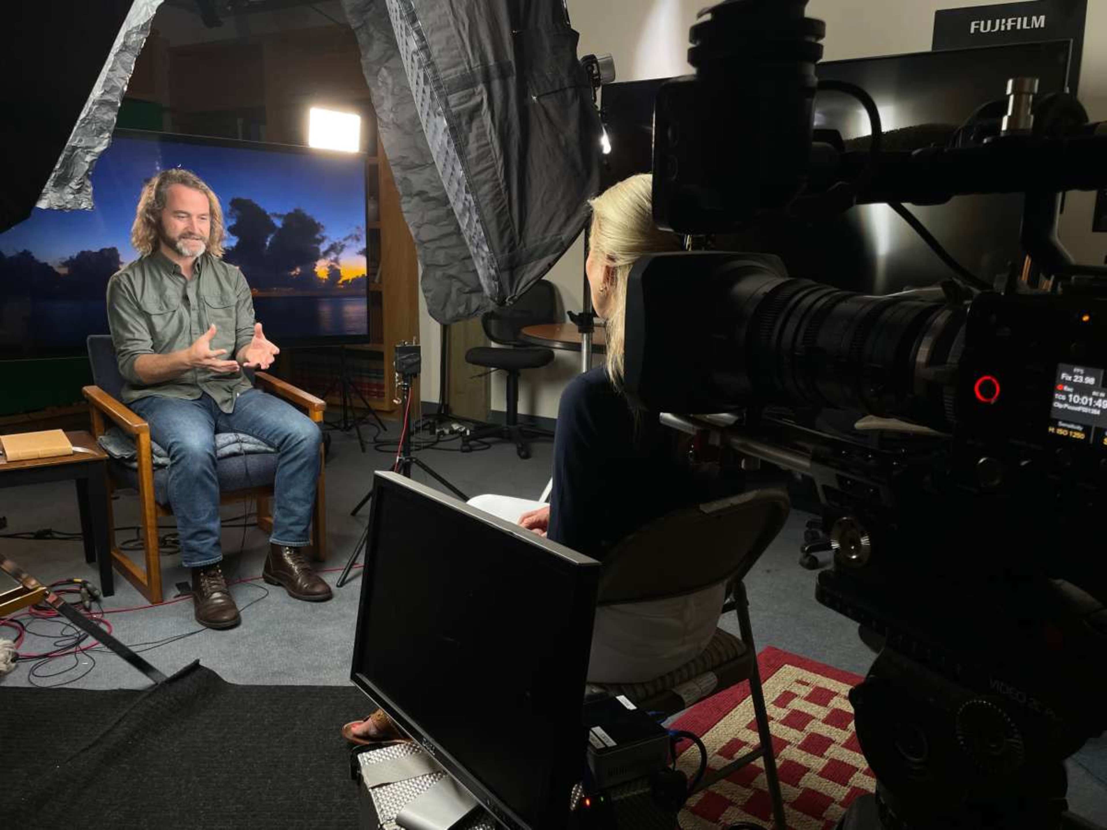 A man with long hair is sitting in a chair and speaking to a woman who is seated across from him, while a camera and lighting setup is positioned nearby.