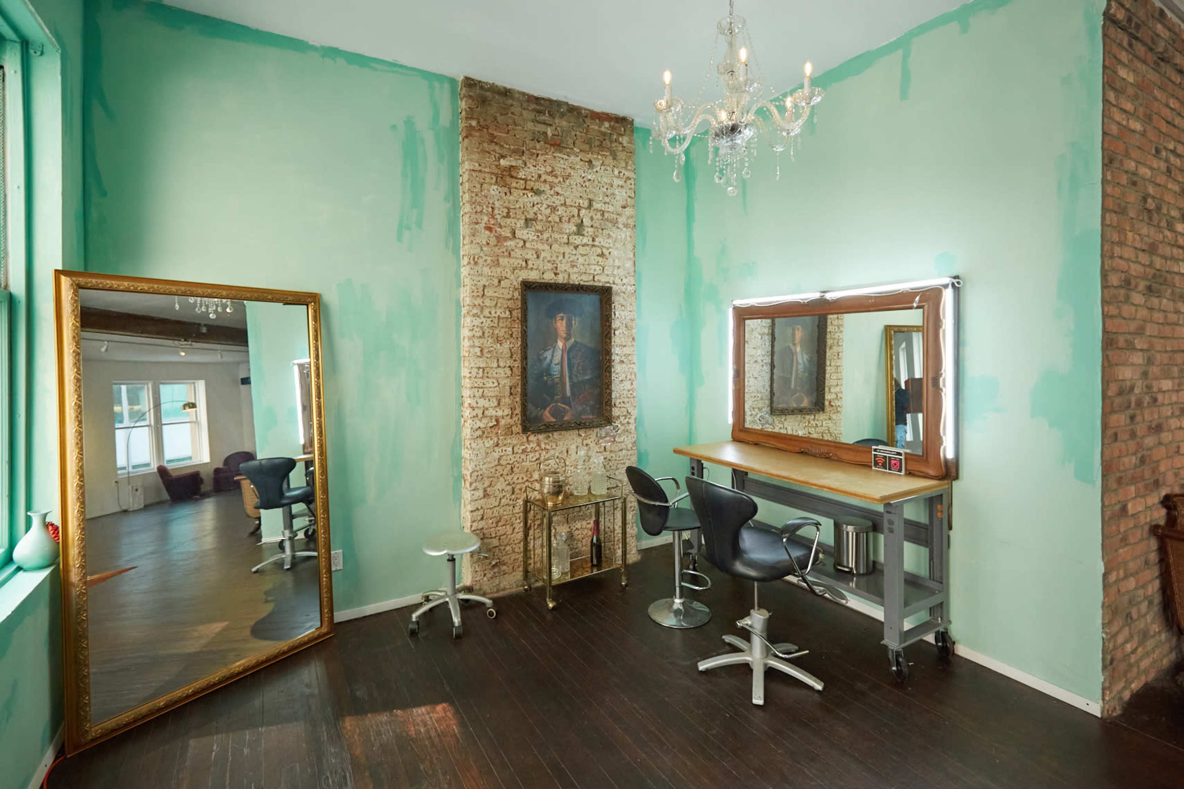 A hair salon with a large mirror and styling station, exposed brick wall, and a chandelier, all set against mint green walls.