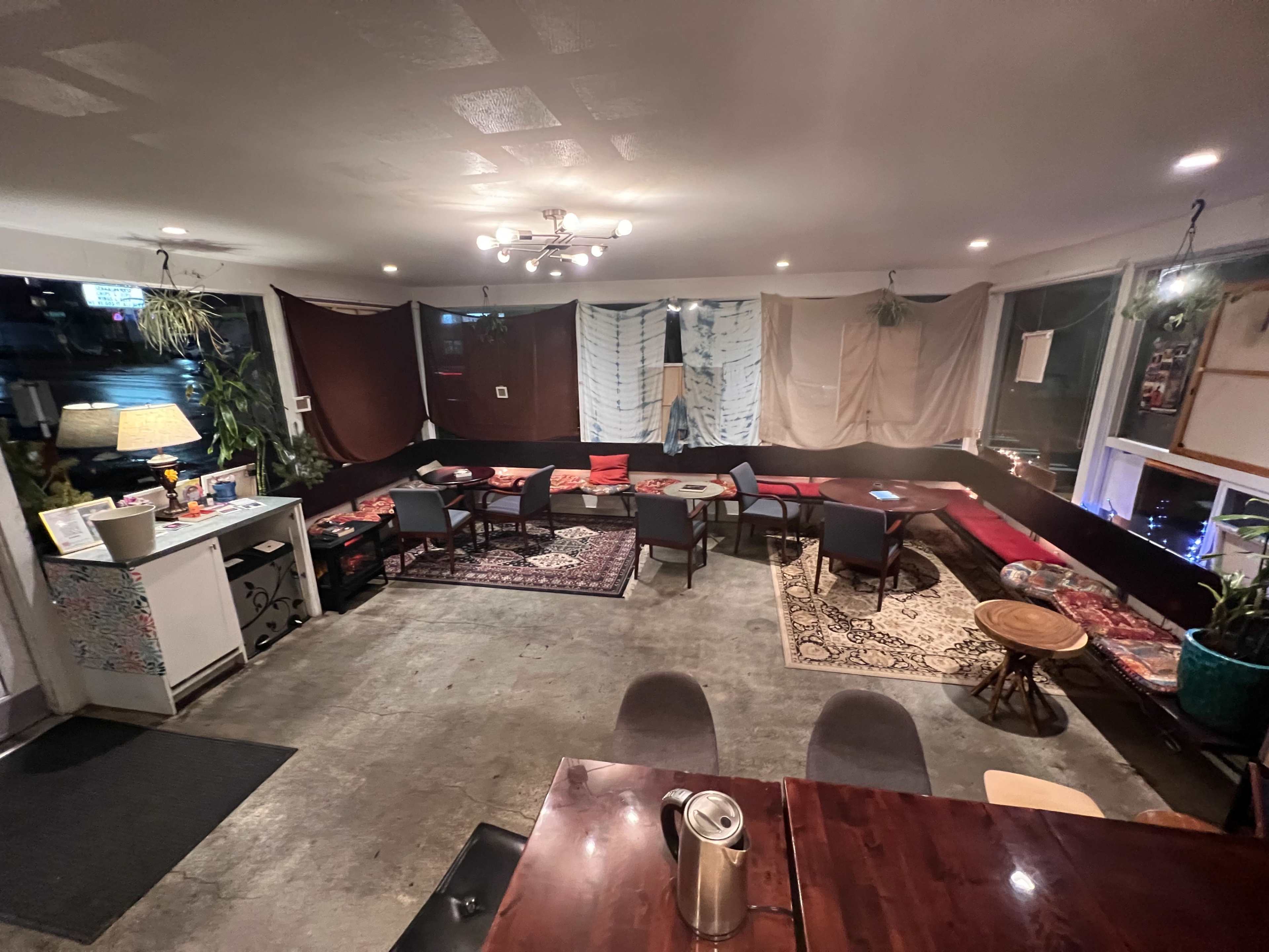 The image shows a cozy interior of a café featuring a low seating area with colorful rugs and cushions, surrounded by plants and soft lighting.