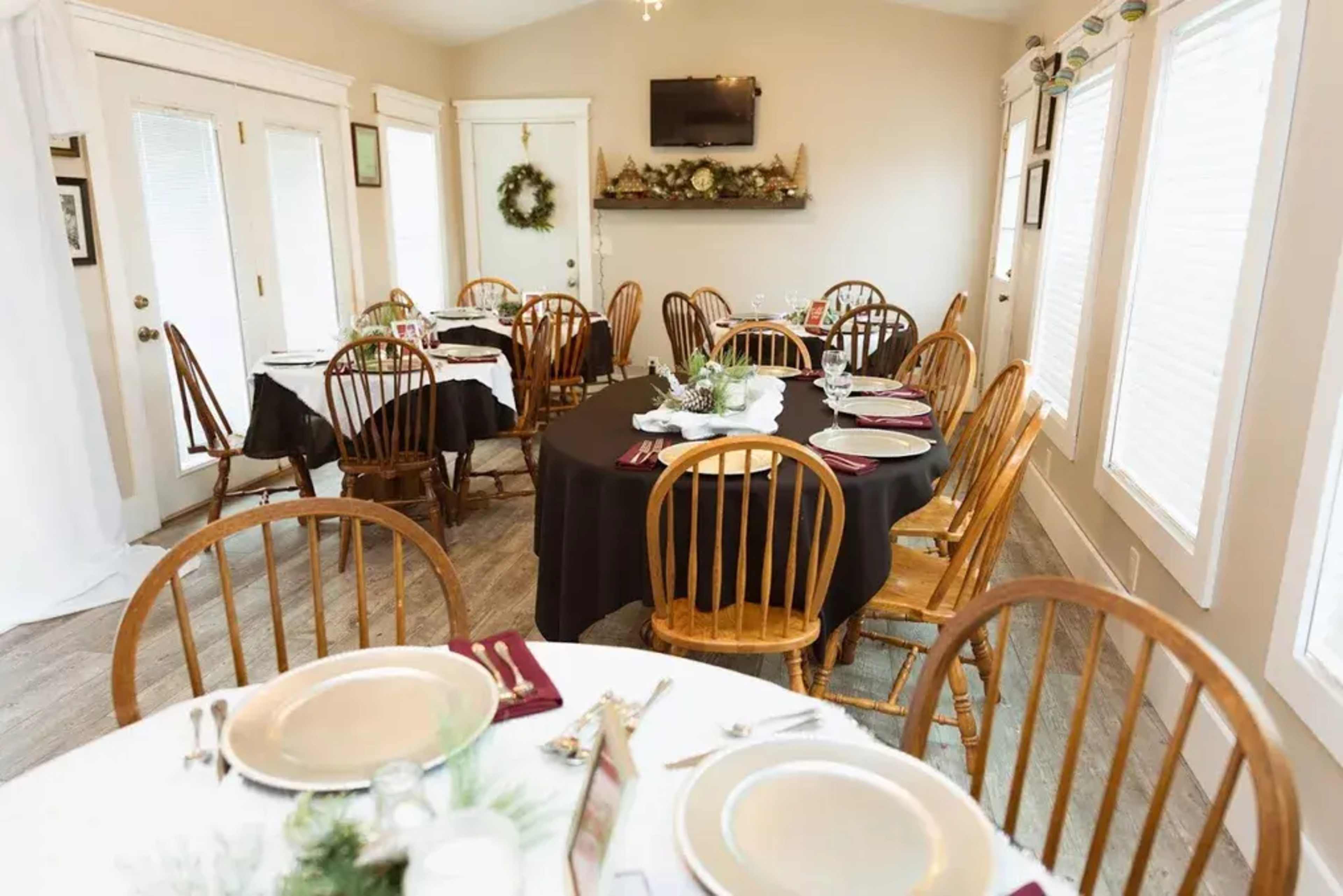 A dining room is set up with several tables, some covered in white tablecloths and others in black, surrounded by wooden chairs and decorated with centerpieces.