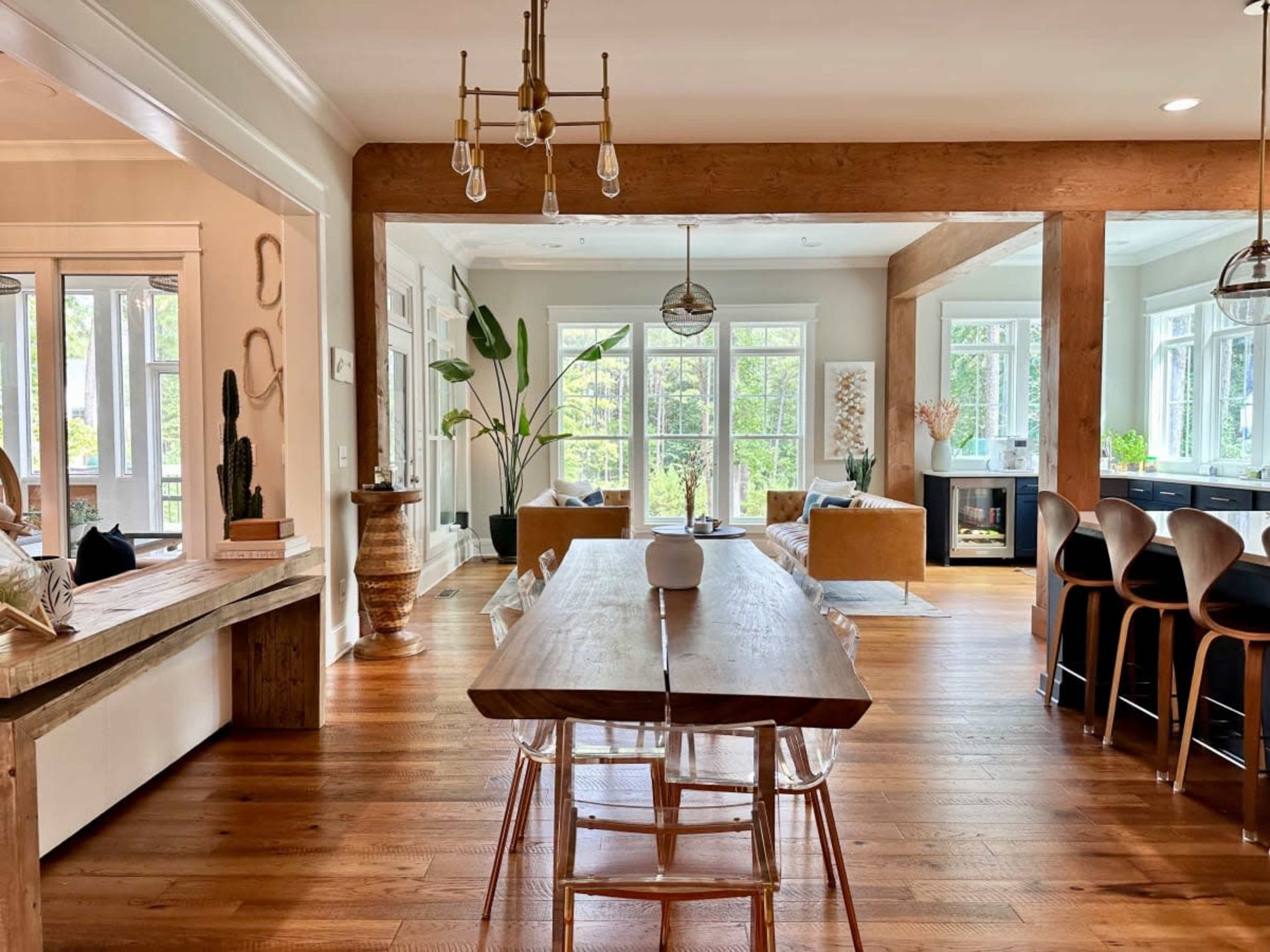 A spacious dining area features a large wooden table surrounded by clear chairs, with large windows and a view of greenery visible in the background.