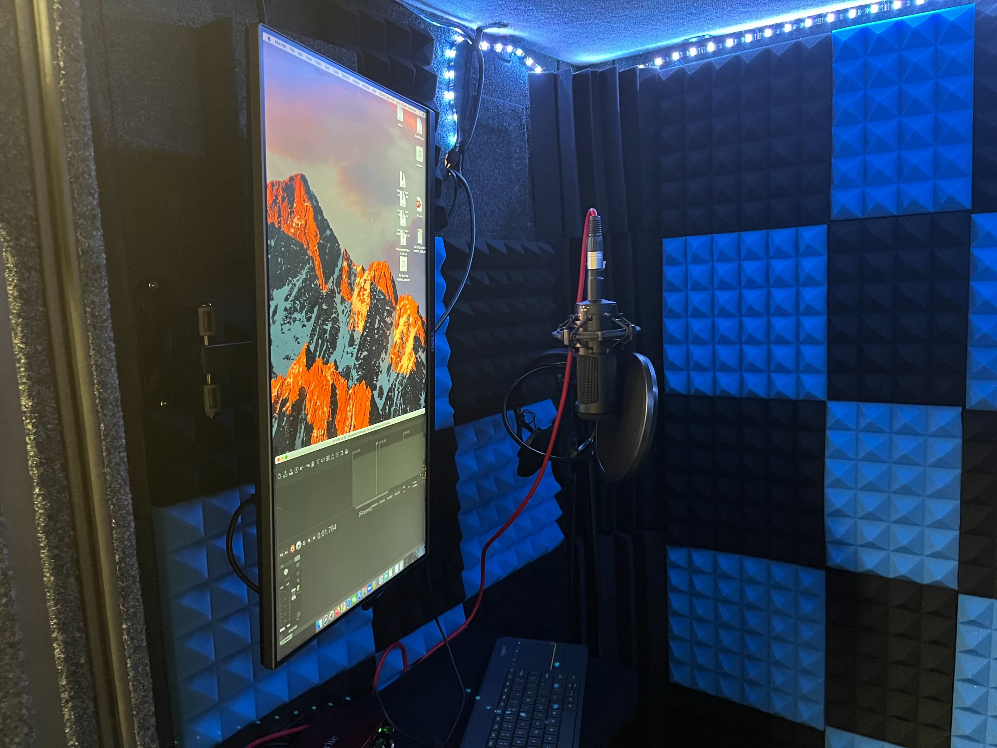 A soundproof recording booth featuring a monitor displaying editing software, a microphone mounted on a stand, and acoustic foam panels in blue and black.