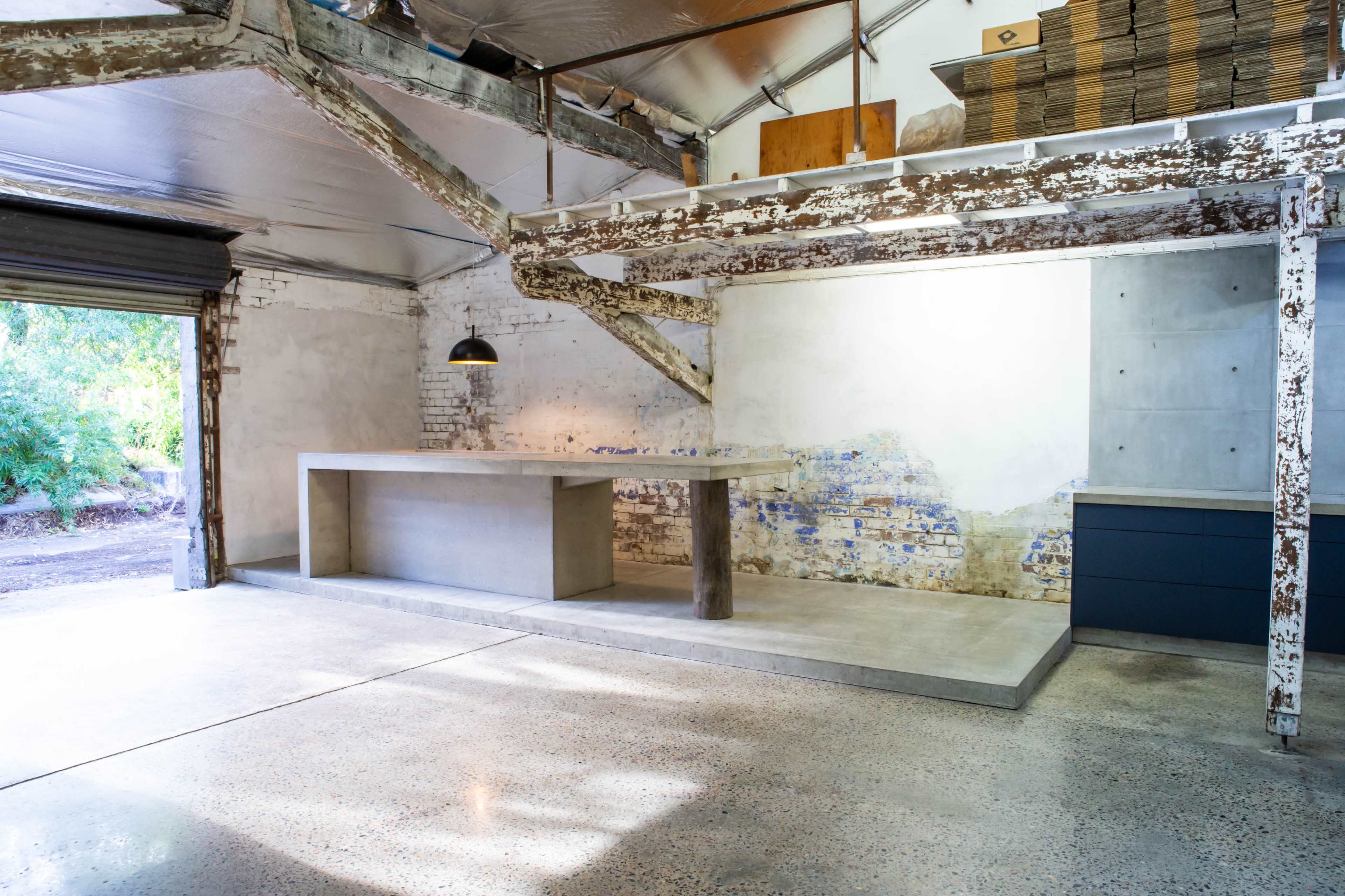 The image shows a modern industrial-style interior with a polished concrete countertop, a large open space featuring exposed wooden beams, and a minimalistic kitchen area with dark cabinetry.