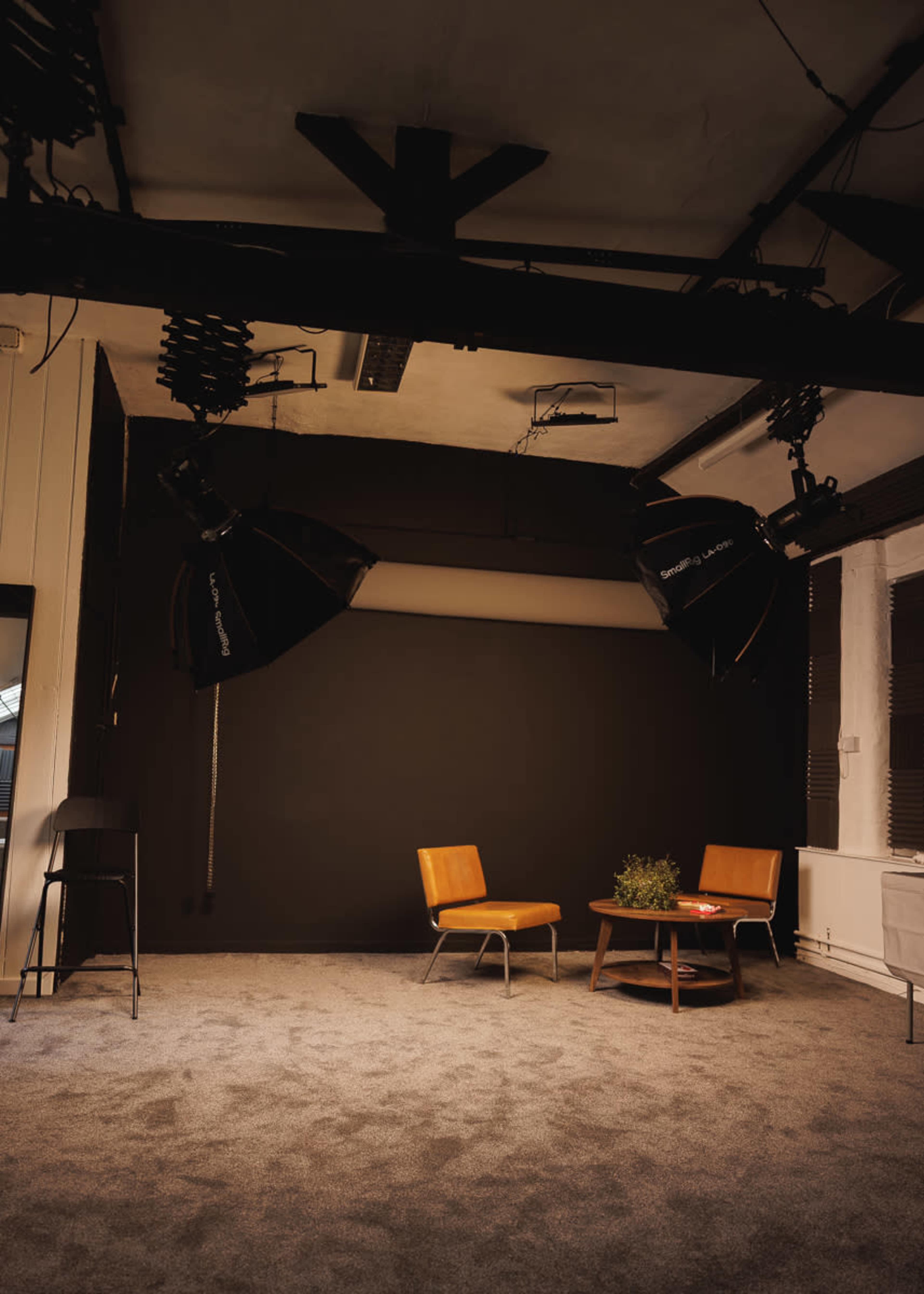 The image shows a minimalist studio setup featuring two wooden chairs, a round table with a potted plant, and overhead lighting against a dark backdrop.