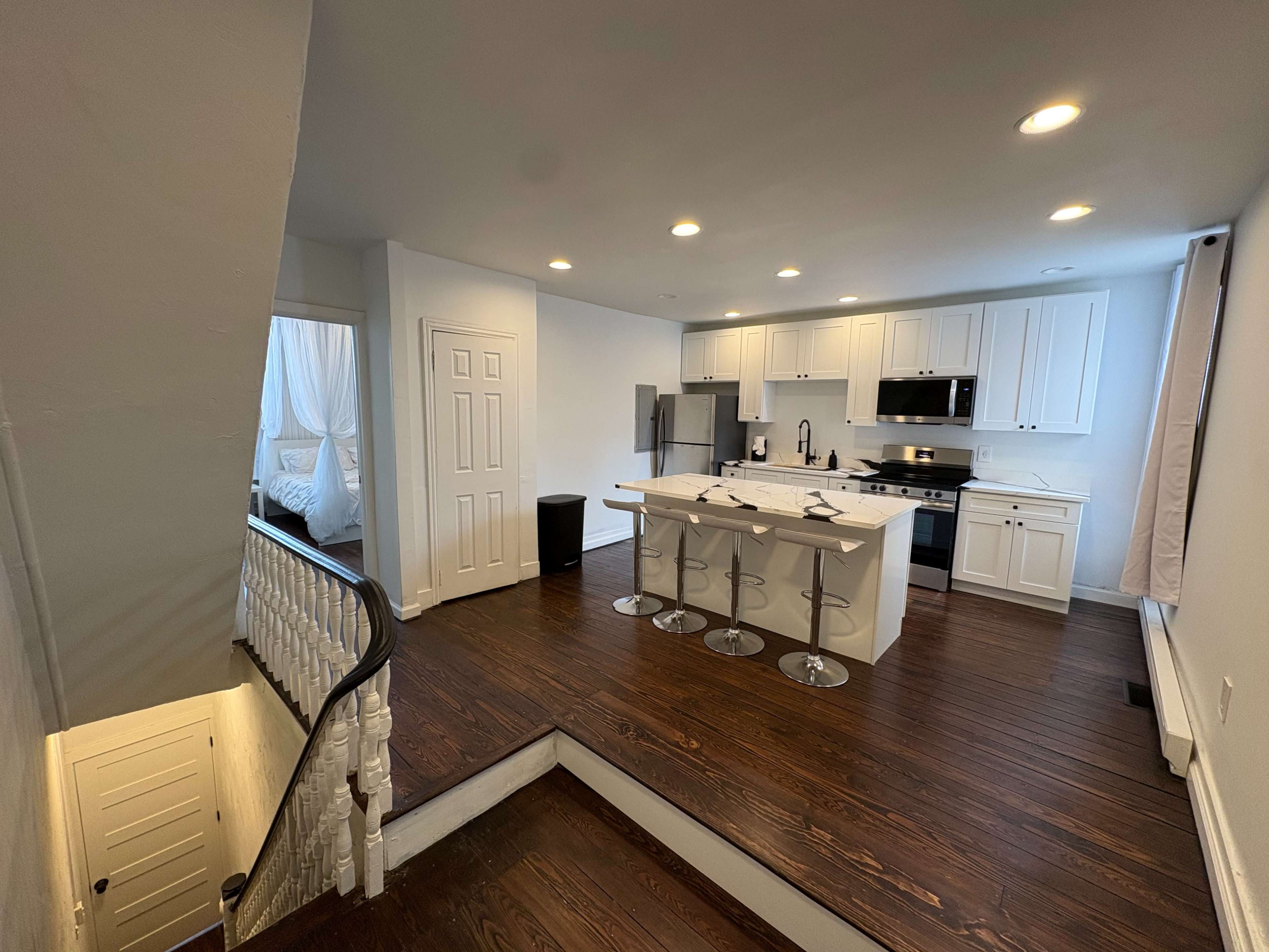 The image shows a modern kitchen with white cabinetry, stainless steel appliances, and a staircase leading to an upper level in a well-lit open space.