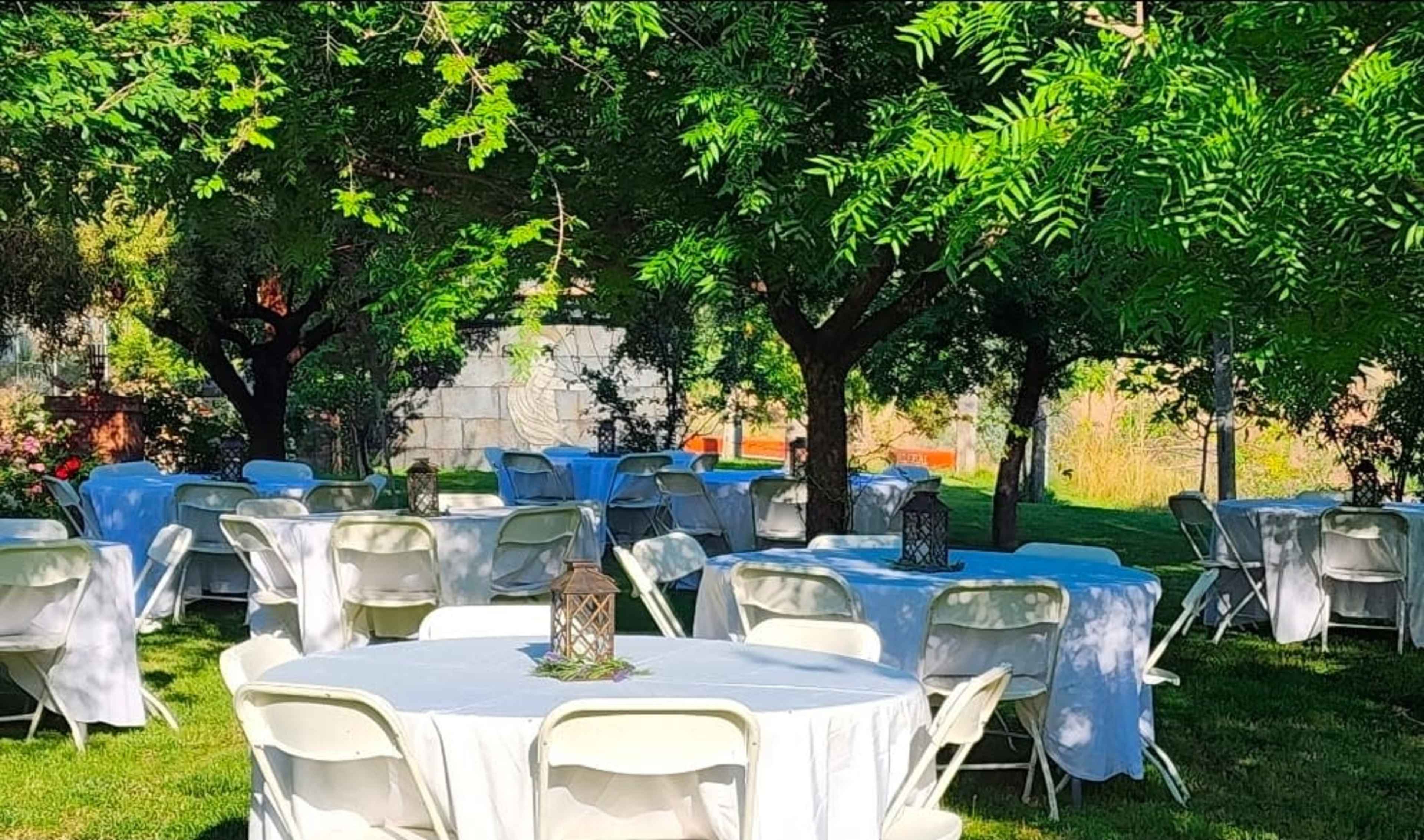 The image shows a garden setting with several round tables covered in white tablecloths, surrounded by lush trees and greenery.