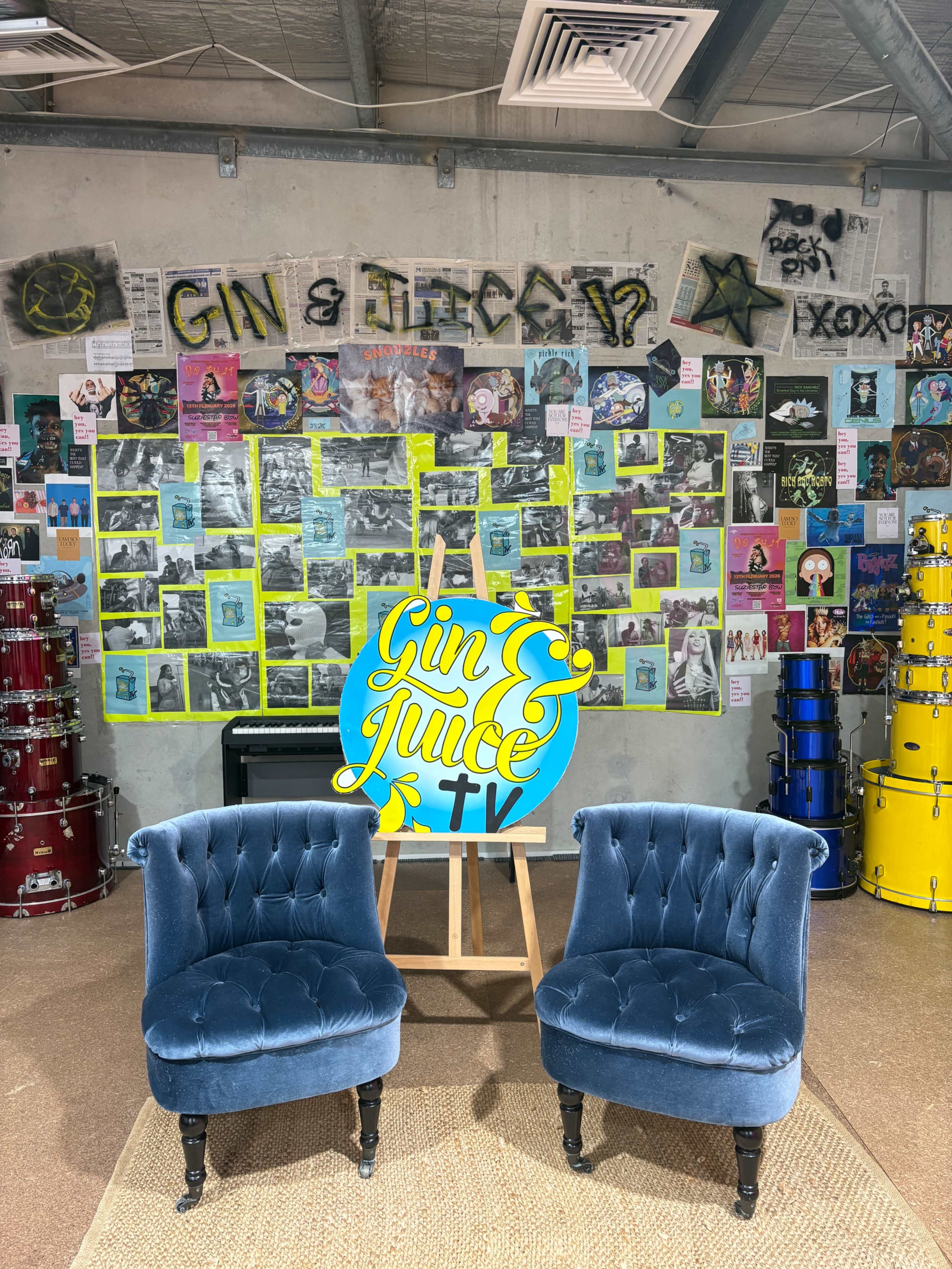 The image shows two blue velvet chairs positioned in front of a colorful wall covered with various posters and a sign that reads "Gin & Juice TV."