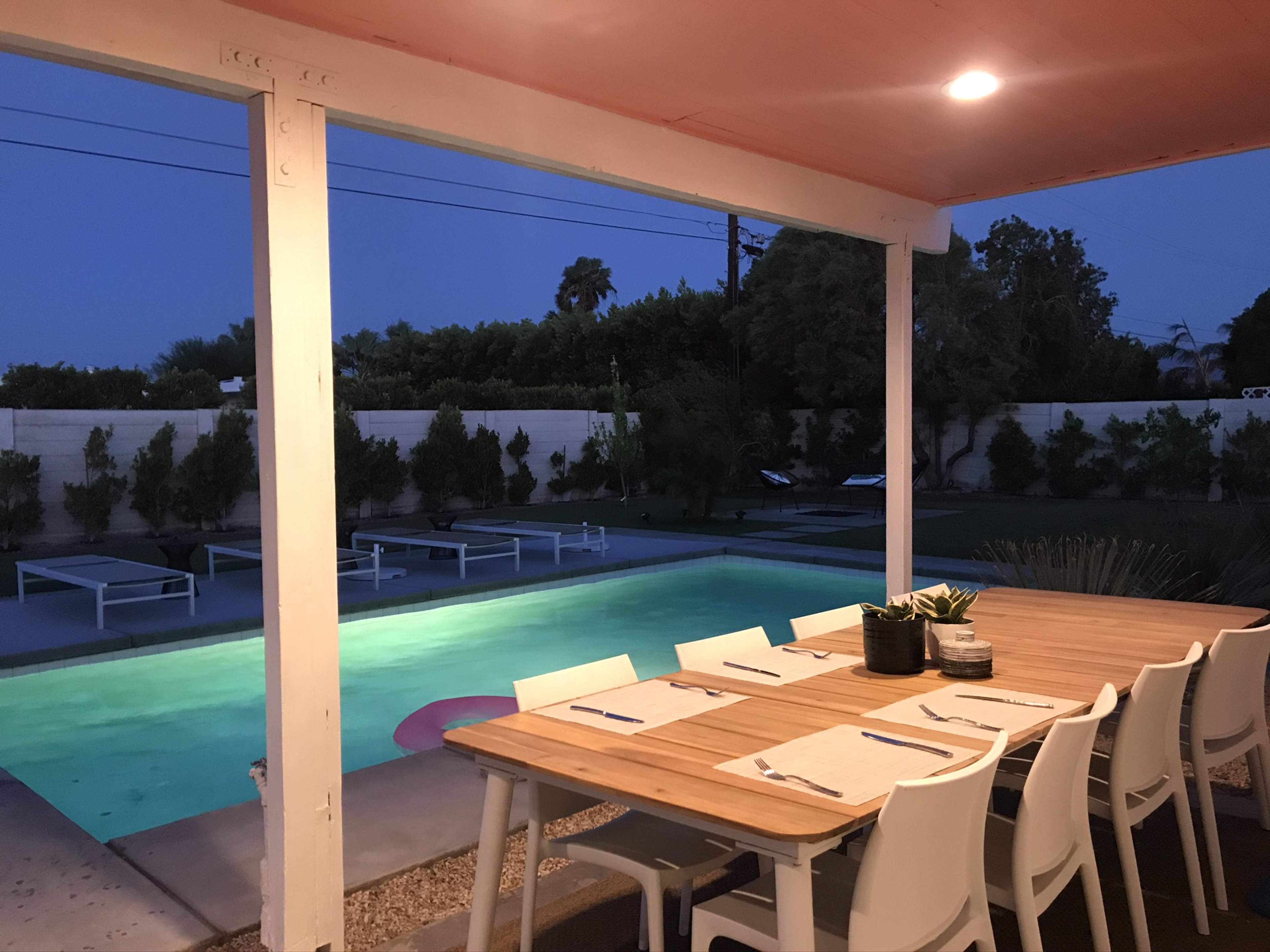 A wooden dining table with white chairs is set up under a covered patio, overlooking a swimming pool illuminated in the evening light.