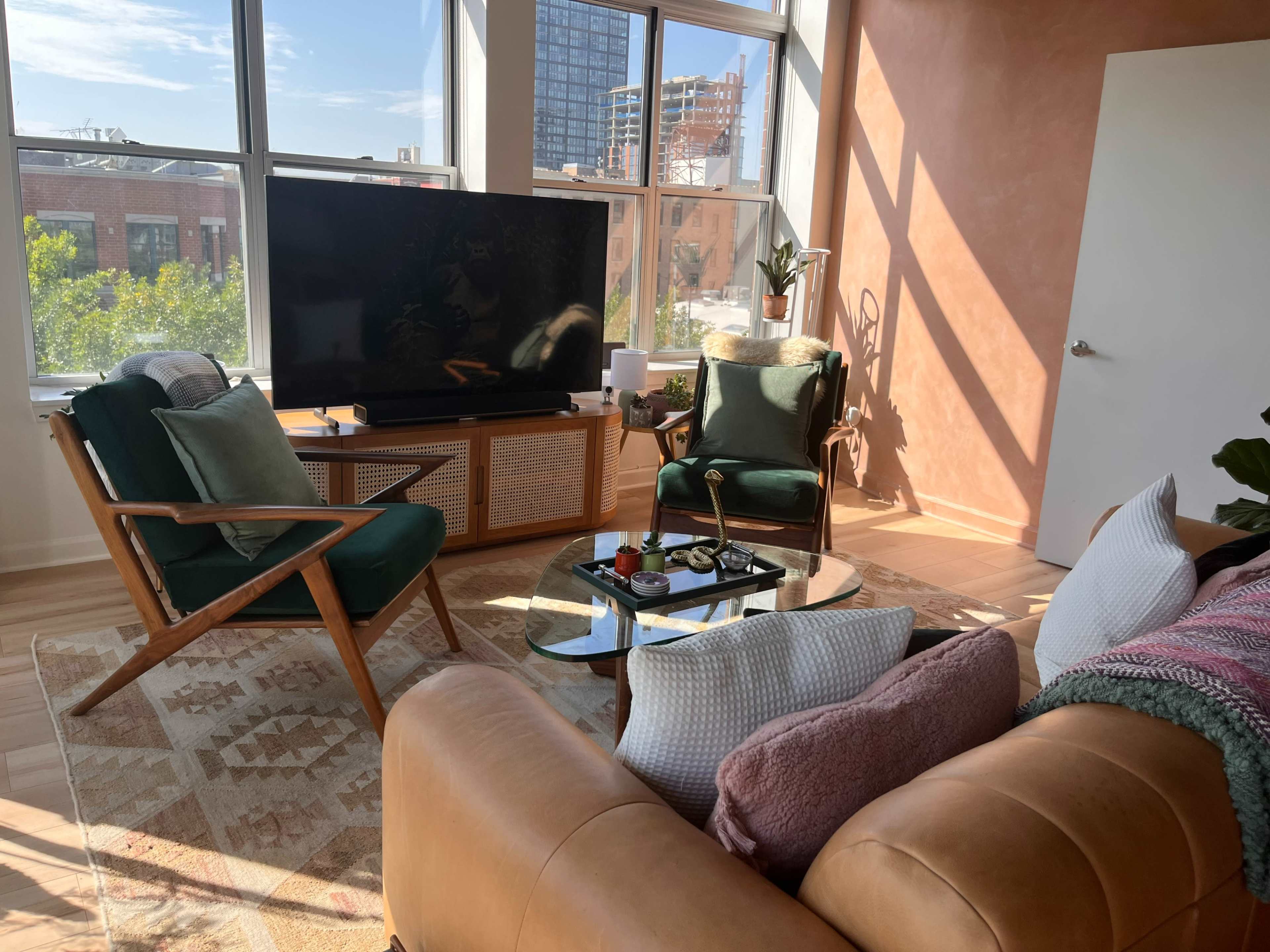 A bright living room features a large television on a wooden stand, two green chairs, a round glass coffee table with decor, and a beige sofa, all set against large windows overlooking a city view.