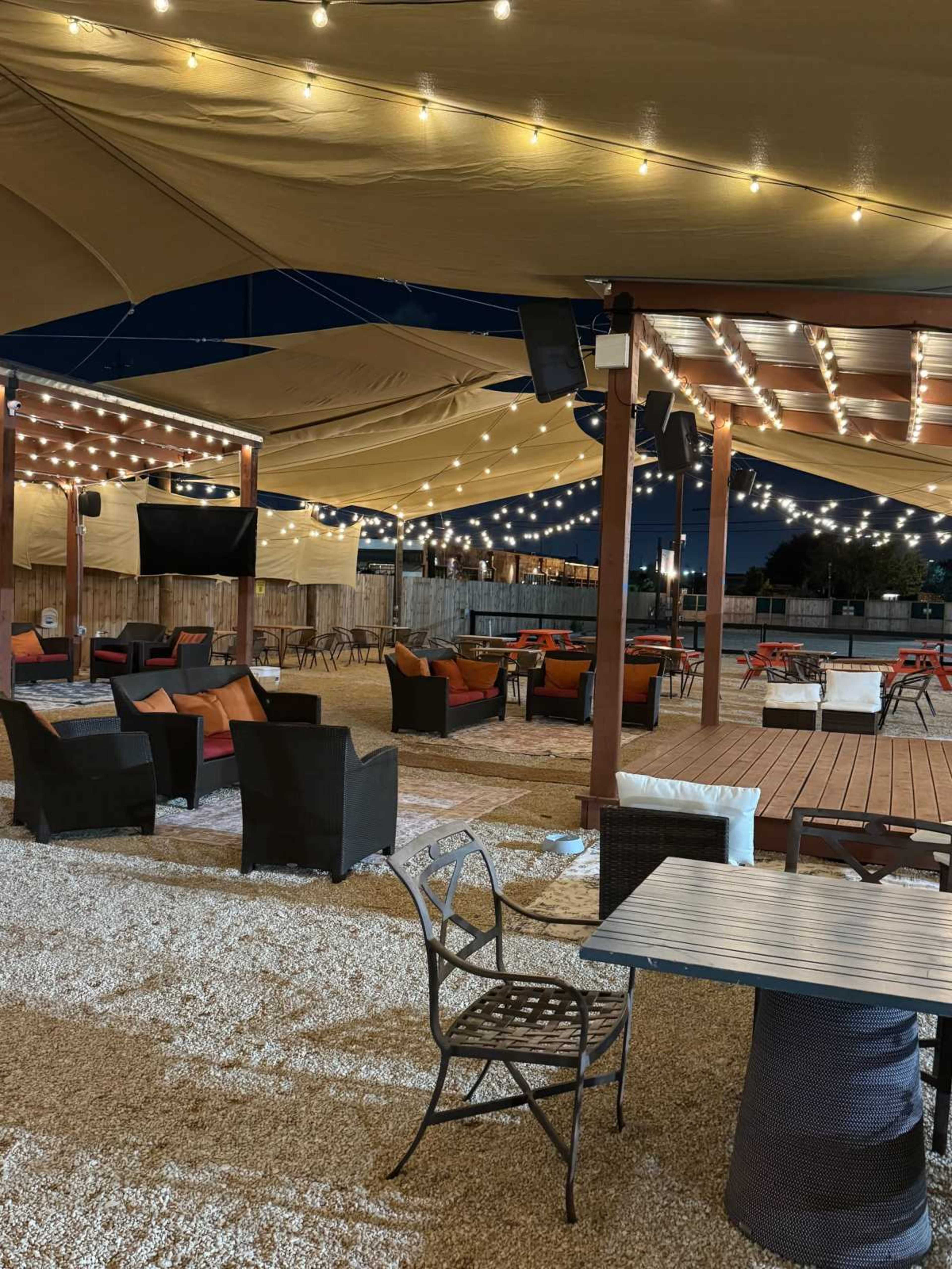 The image shows an outdoor patio area with lounge chairs and tables beneath a large canopy, illuminated by string lights at night.