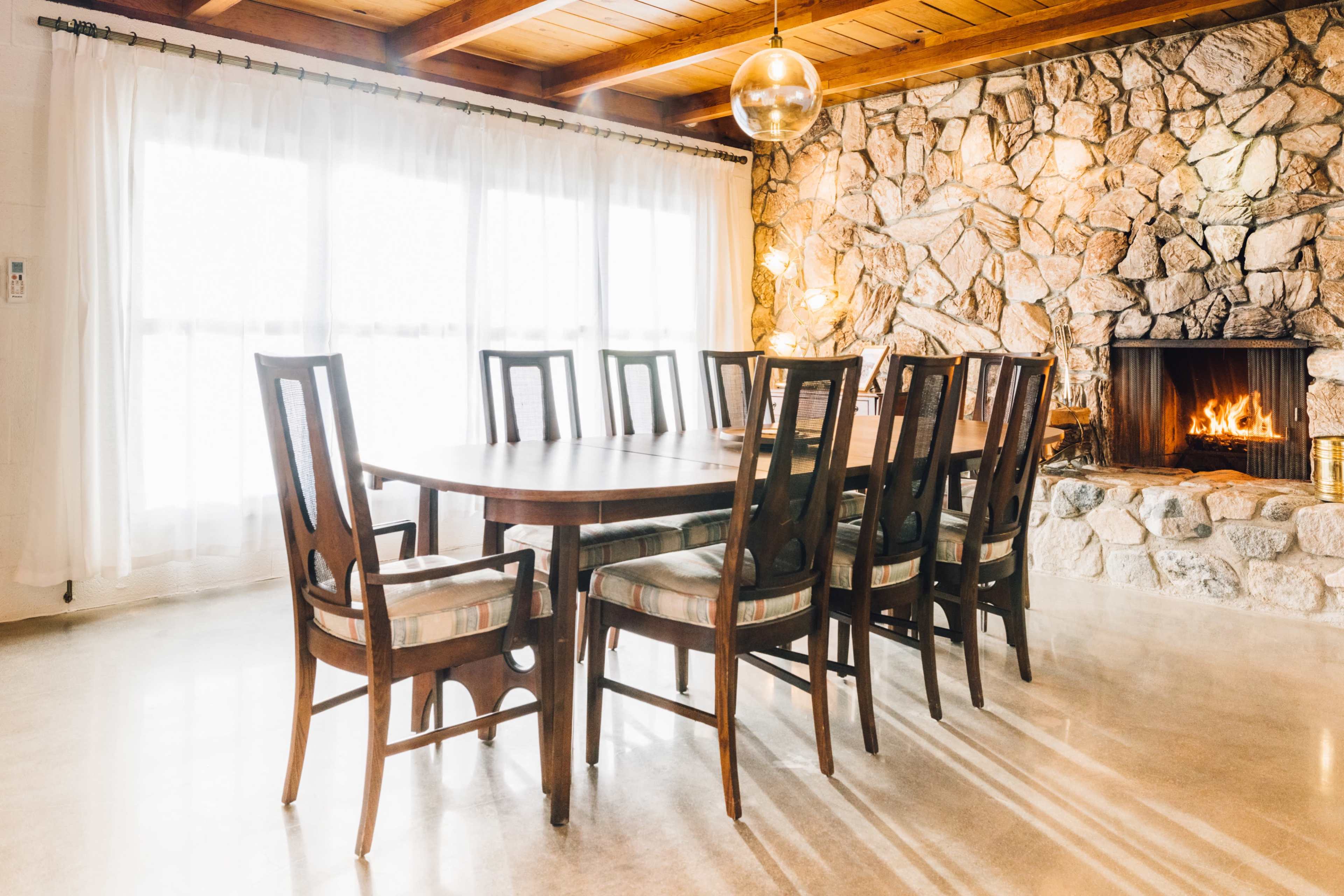 A spacious dining room features a long wooden table surrounded by eight chairs, with a stone fireplace and large windows in the background.