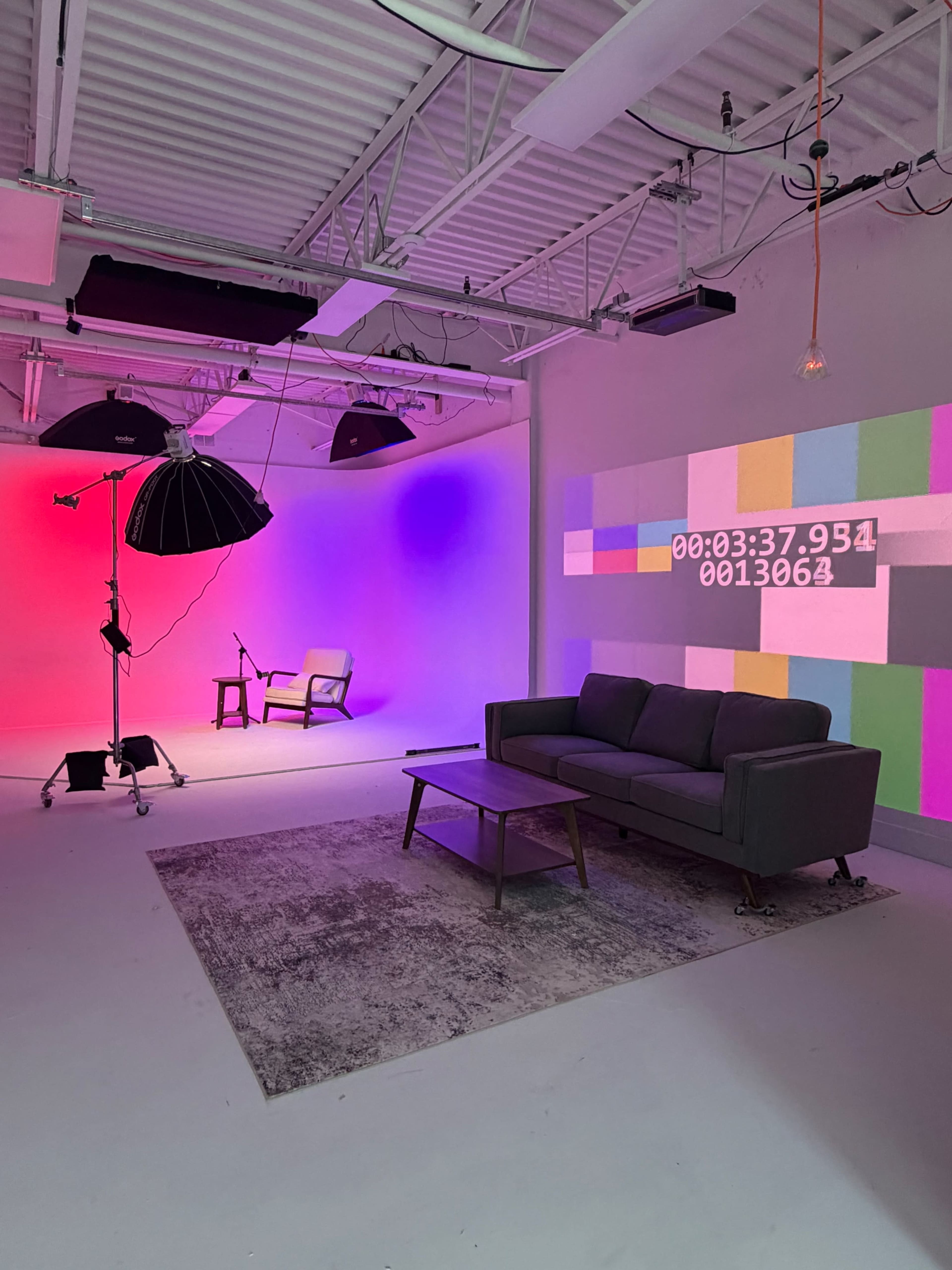 The image shows a brightly lit studio space with a sofa, coffee table, and a large screen displaying colorful stripes and a timer, alongside a photography light setup.