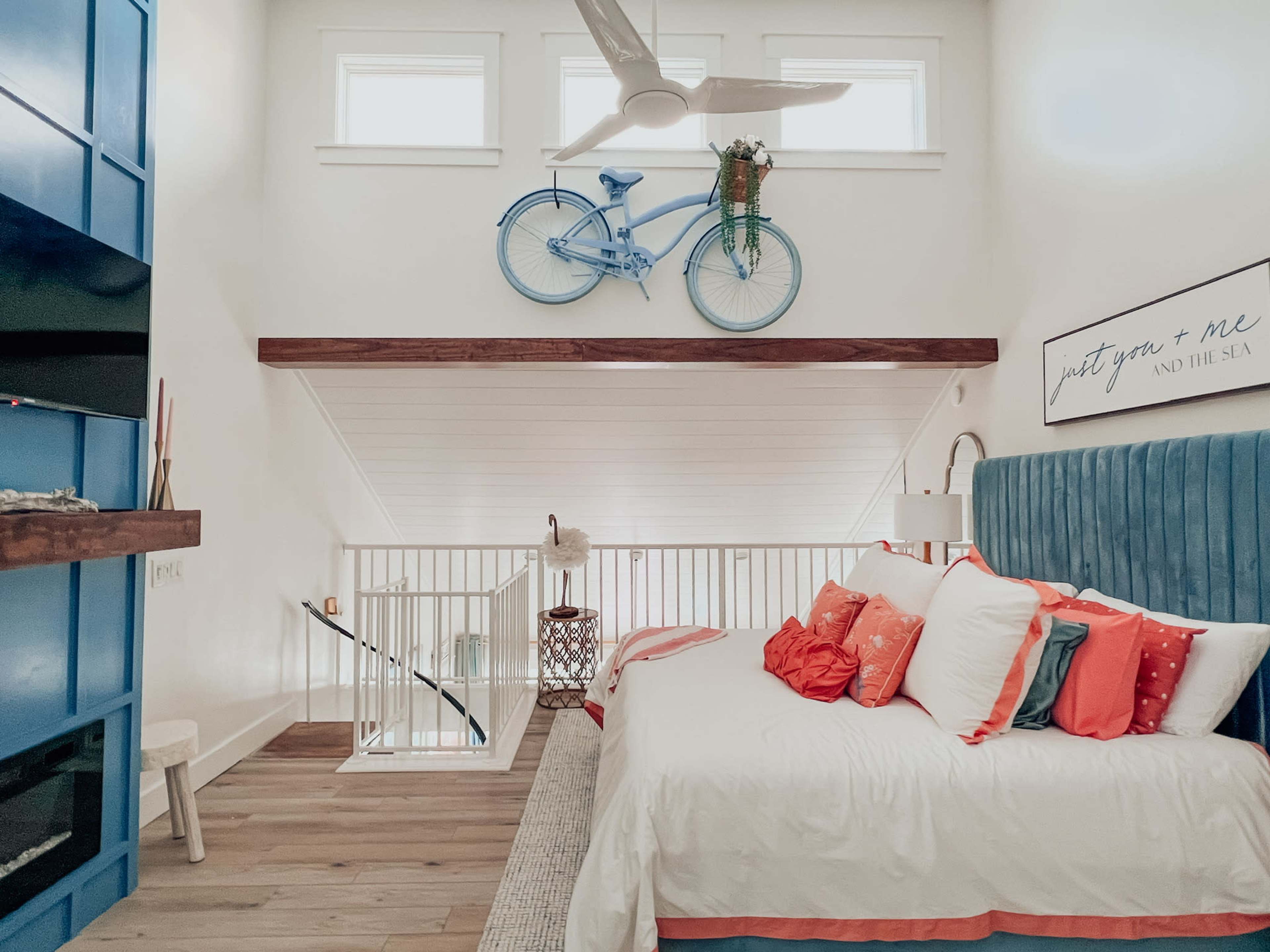 A bright bedroom features a blue bicycle mounted on the wall above a king-sized bed with colorful pillows, alongside a staircase leading to a lofted area.