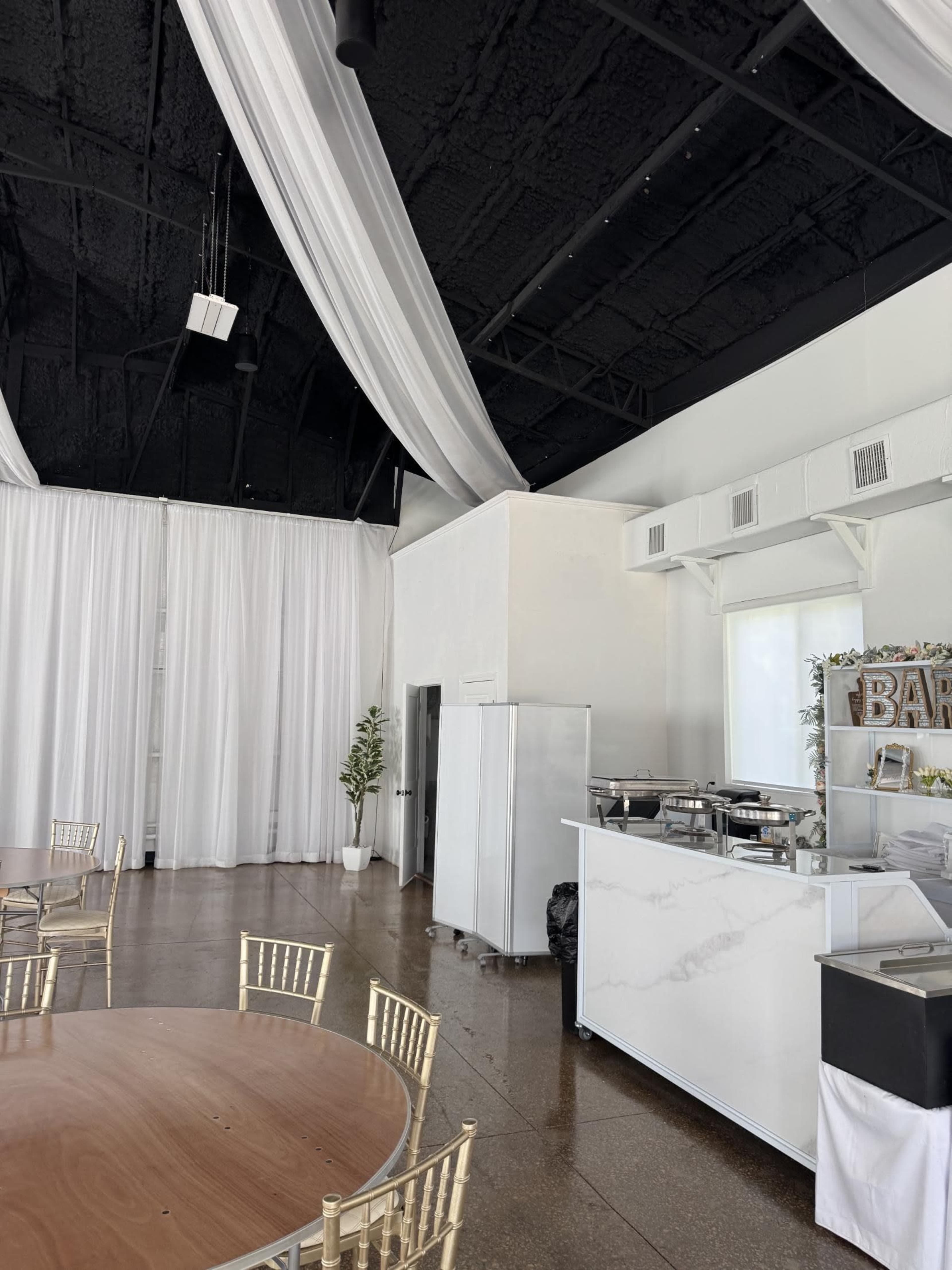 The image shows a spacious indoor venue with white drapery, high black ceilings, and a serving area featuring a silver food station and a refrigerator.