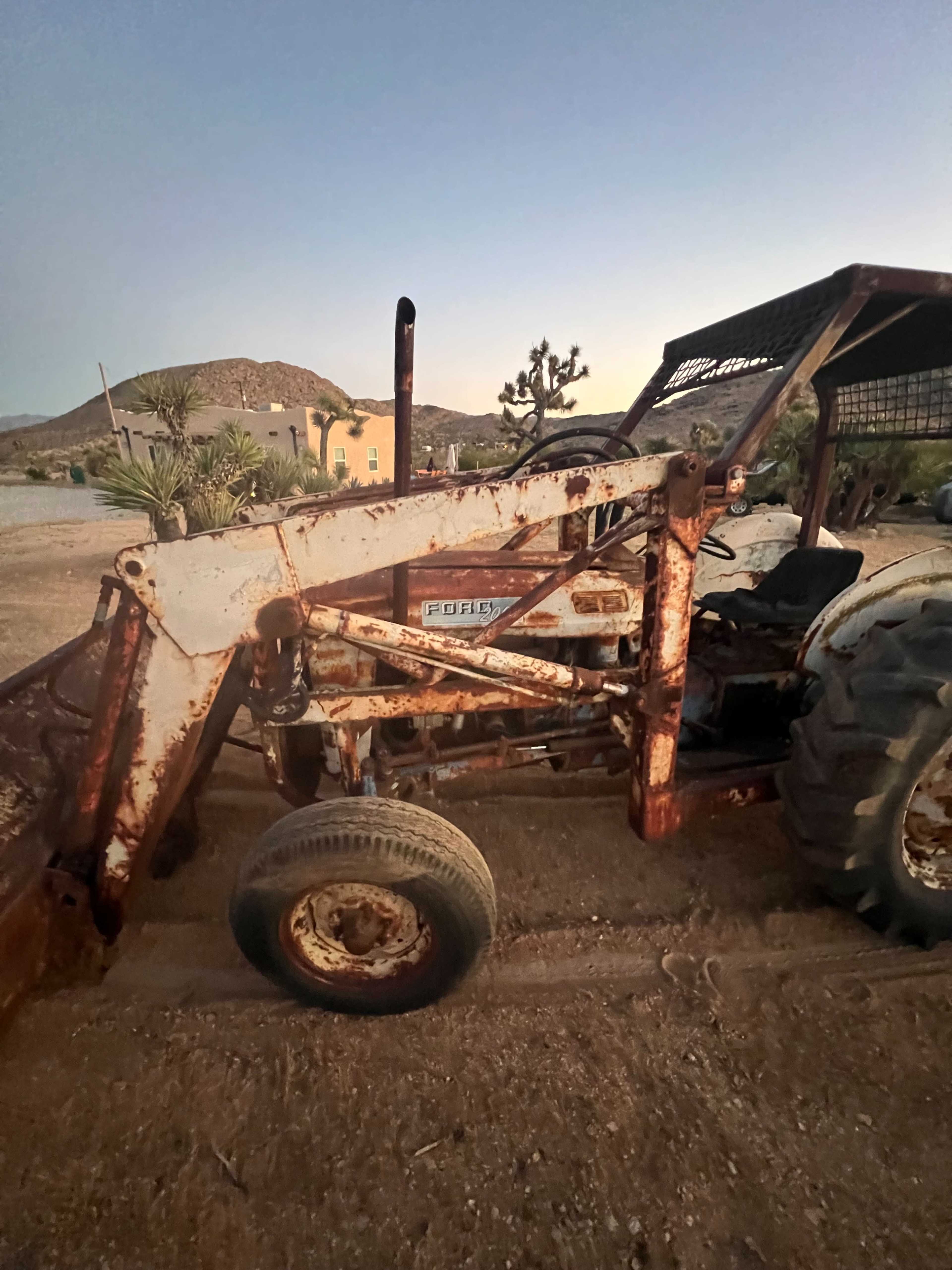 A rusty tractor is parked on a dirt surface with a mountainous landscape in the background.