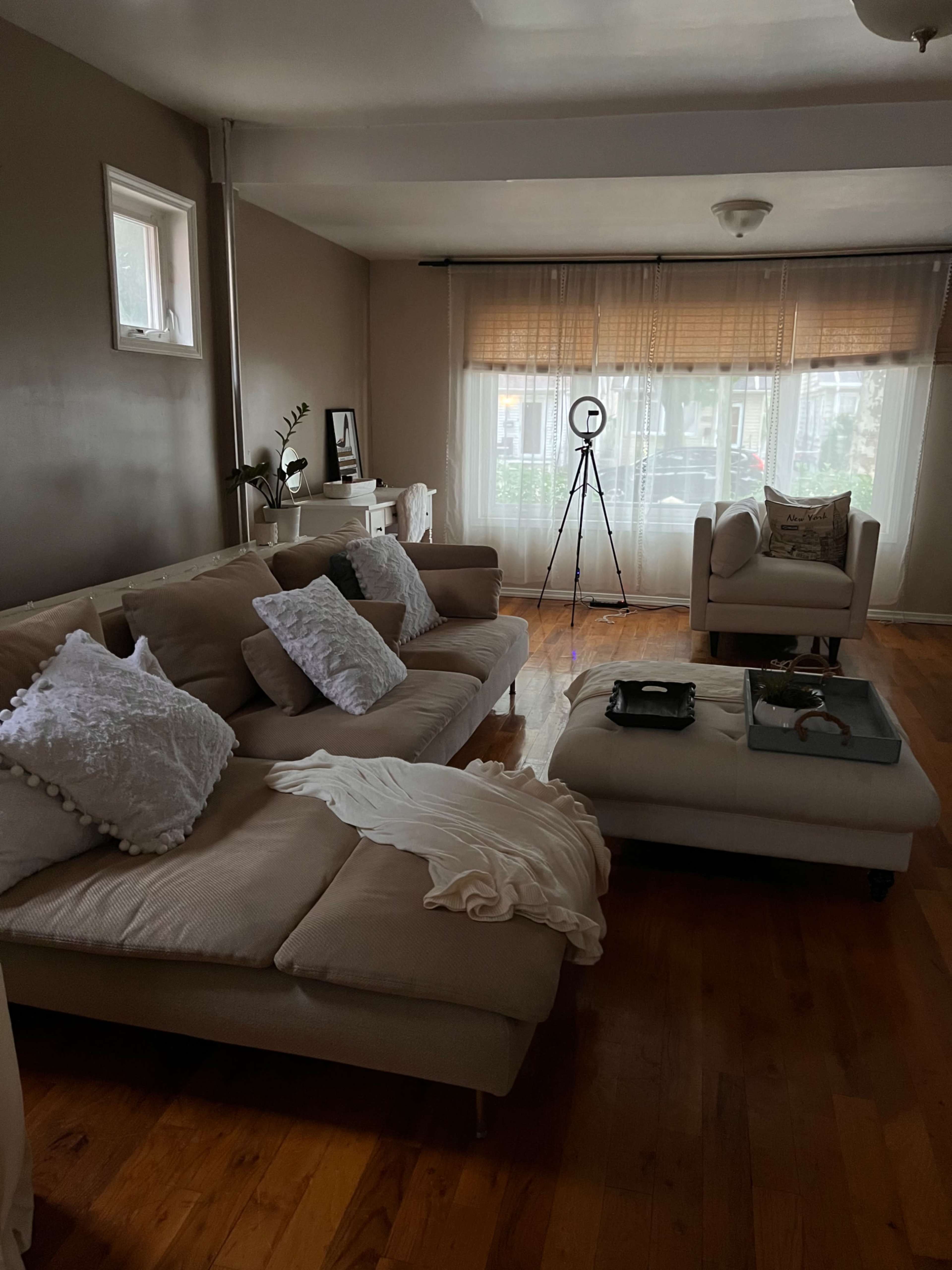 A simple living room with a light-colored sectional sofa, a coffee table, and a large window with sheer curtains.