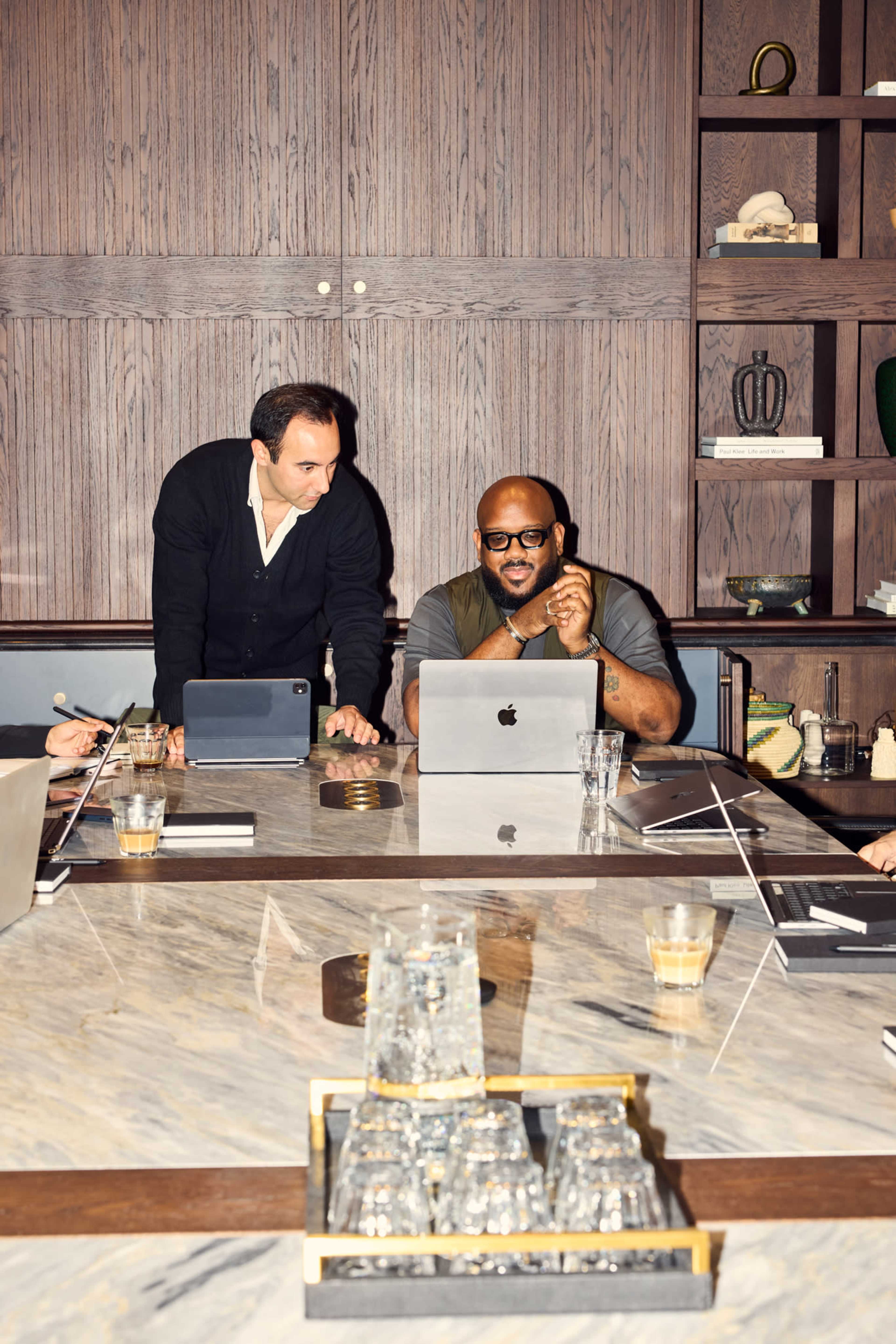 A group of professionals is engaged in a discussion around a conference table, with one person standing while others sit, all focused on laptops and documents.