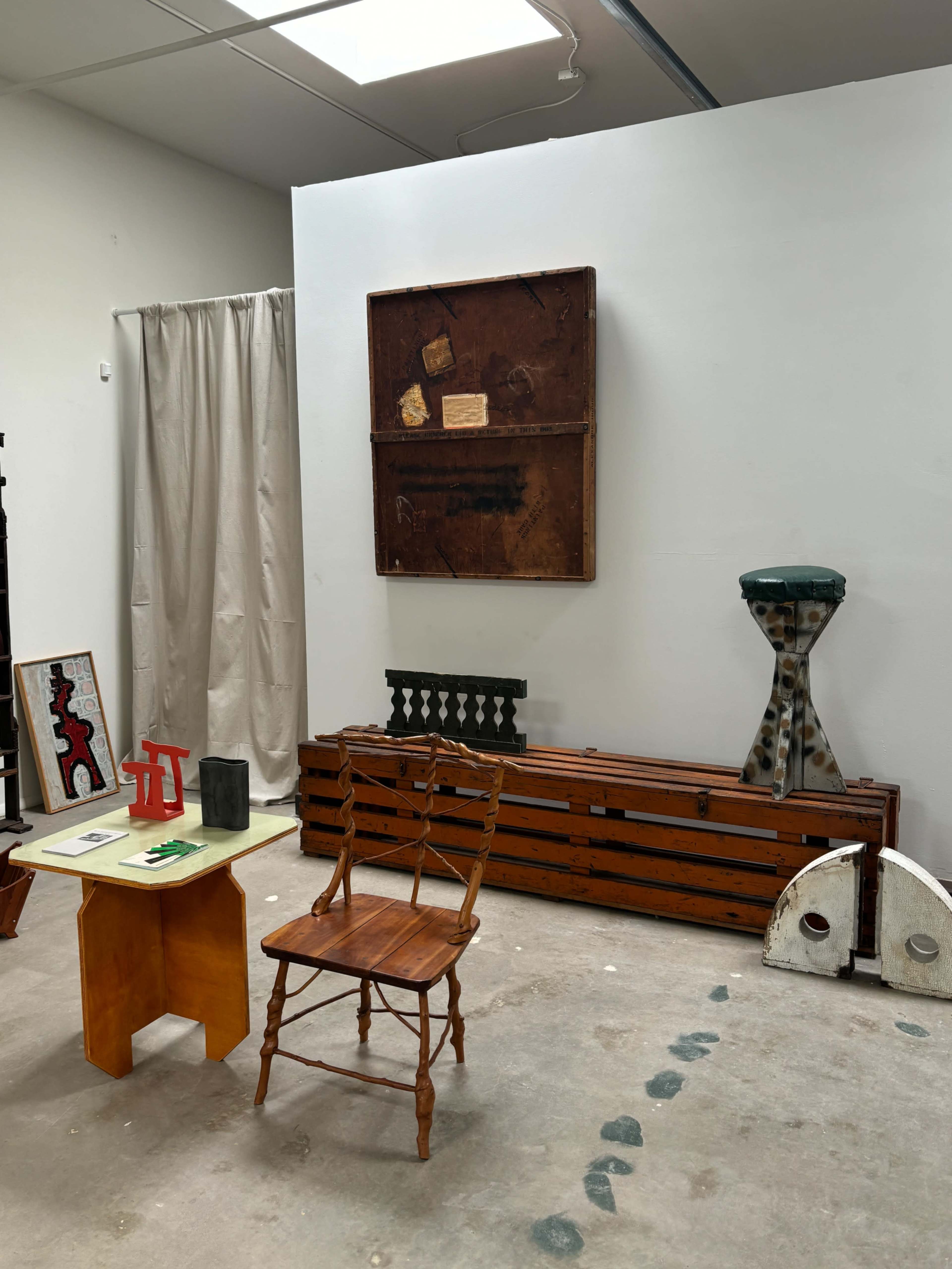 The image shows an art studio featuring a wooden chair, a small table with a green top, various wooden pieces along the wall, and a large framed item hanging on the white wall.