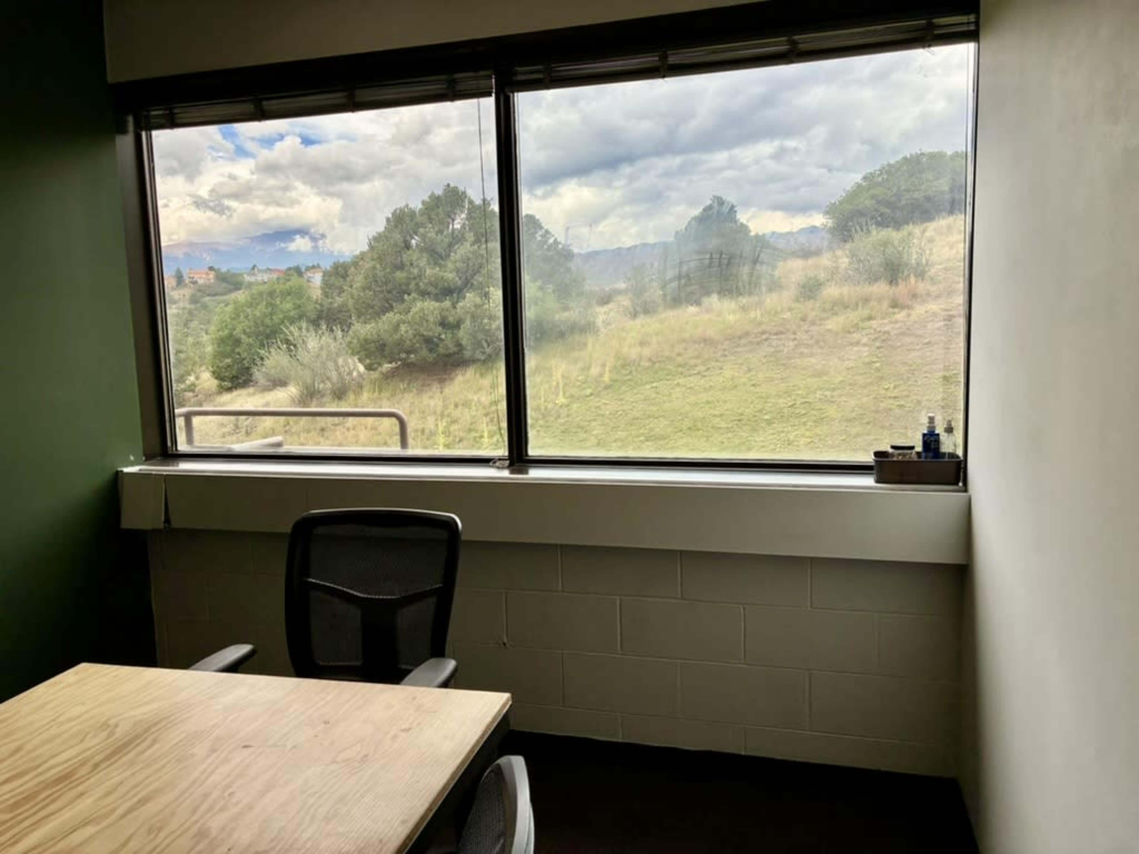 A small office features a wooden table and an empty chair, with a large window revealing a view of grassy hills and cloudy skies outside.