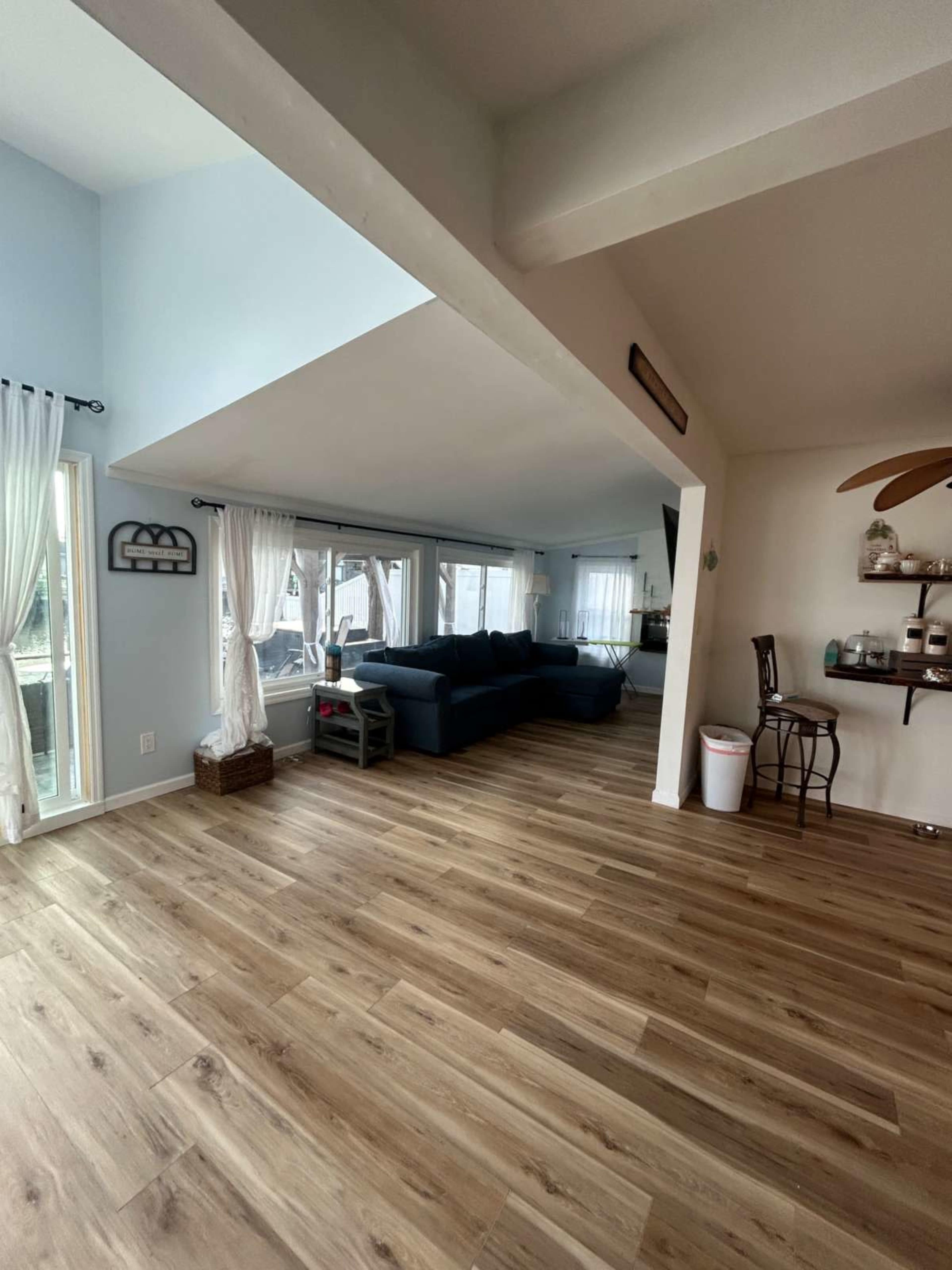 The image shows a spacious living area with light blue walls, a dark blue sofa, large windows, and wooden flooring.