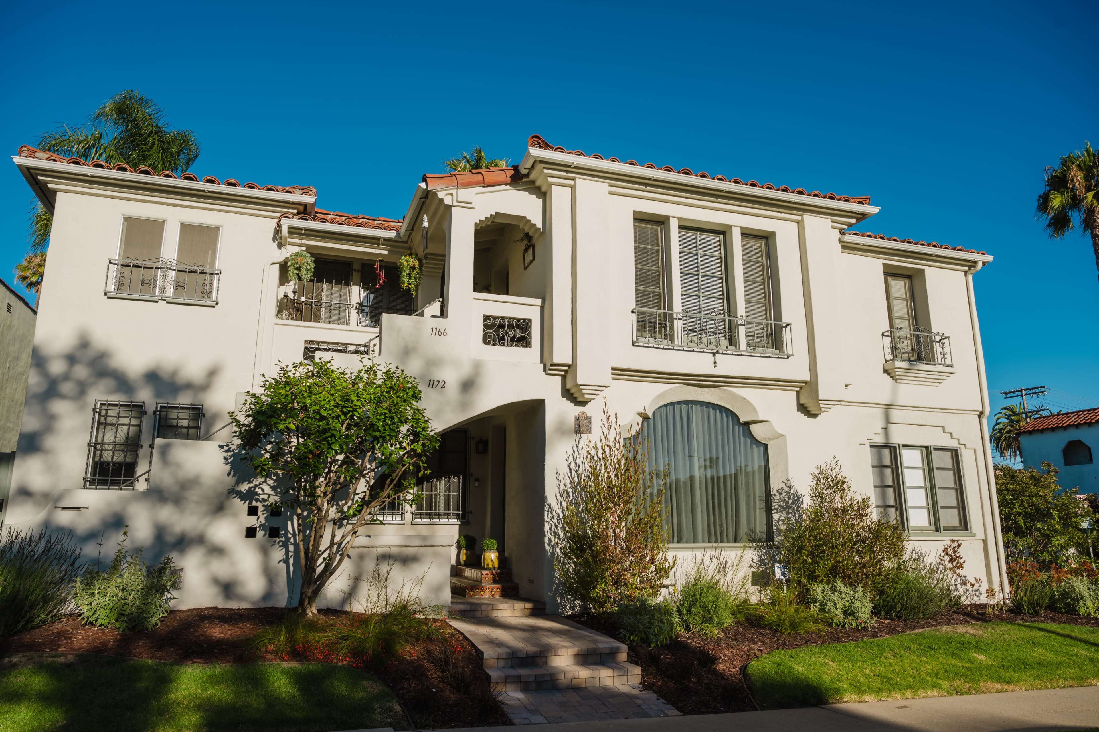 The image shows a two-story white apartment building with a tiled roof, decorative ironwork on the windows, and landscaped greenery in front.