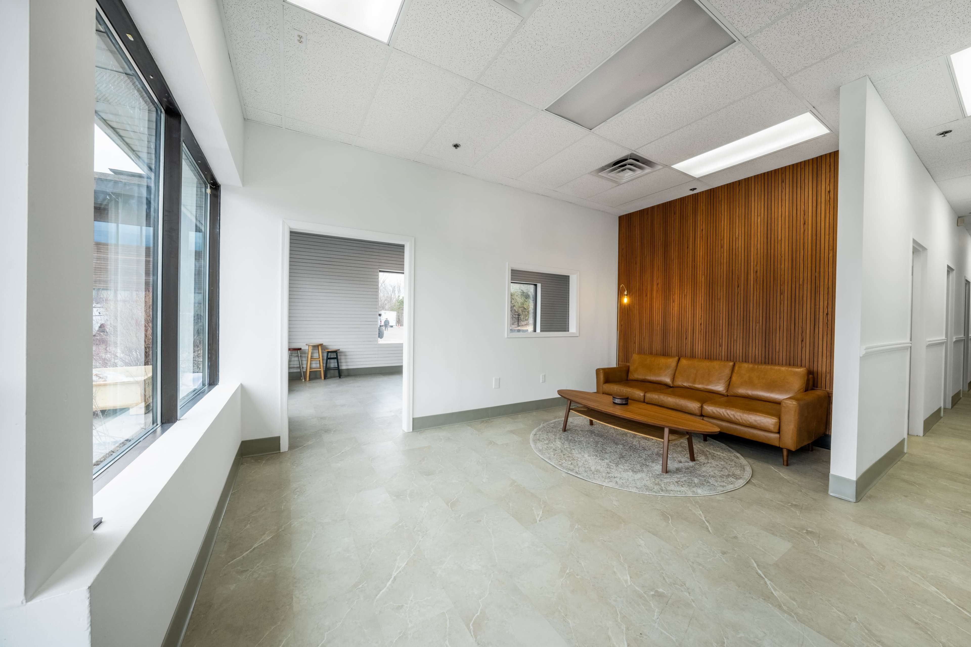 The image shows a modern waiting area featuring a brown leather sofa, a round rug, and a wood-paneled wall, with large windows allowing natural light.