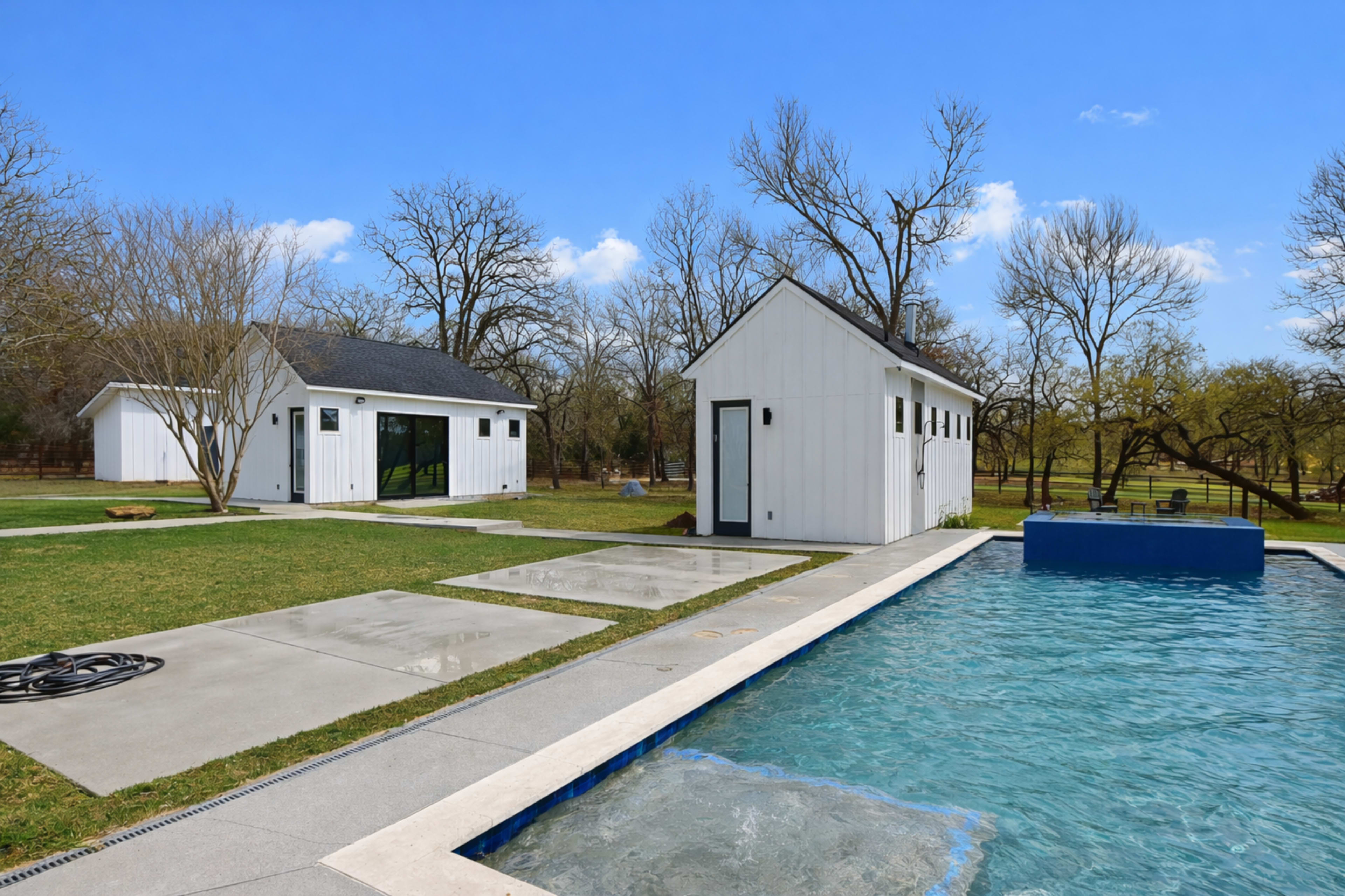 The image shows a modern backyard with a swimming pool surrounded by concrete decking, featuring two white houses in the background and trees in a clear blue sky.