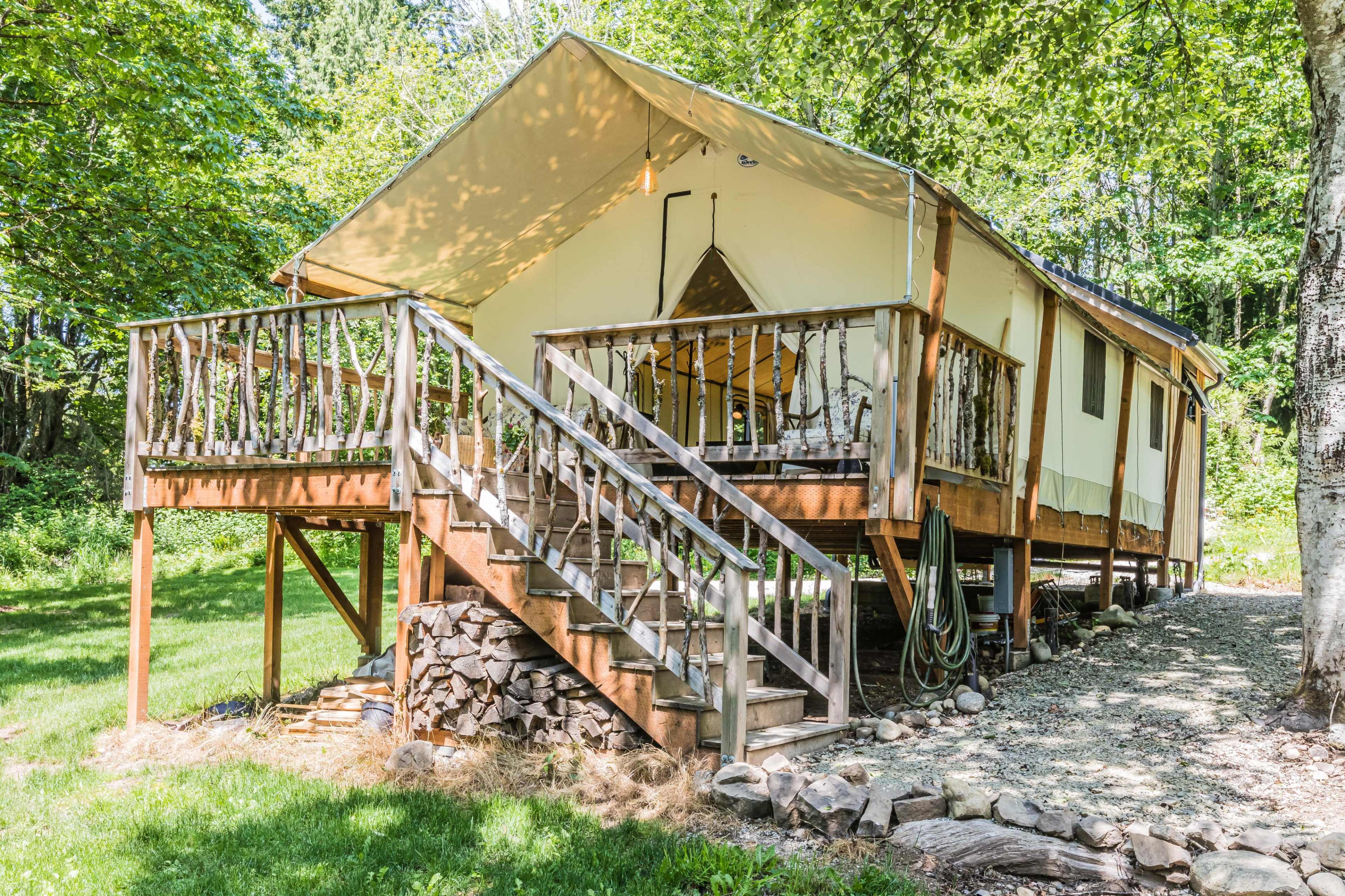 A tent cabin sits elevated on a wooden platform with a set of stairs leading up to a deck surrounded by trees.