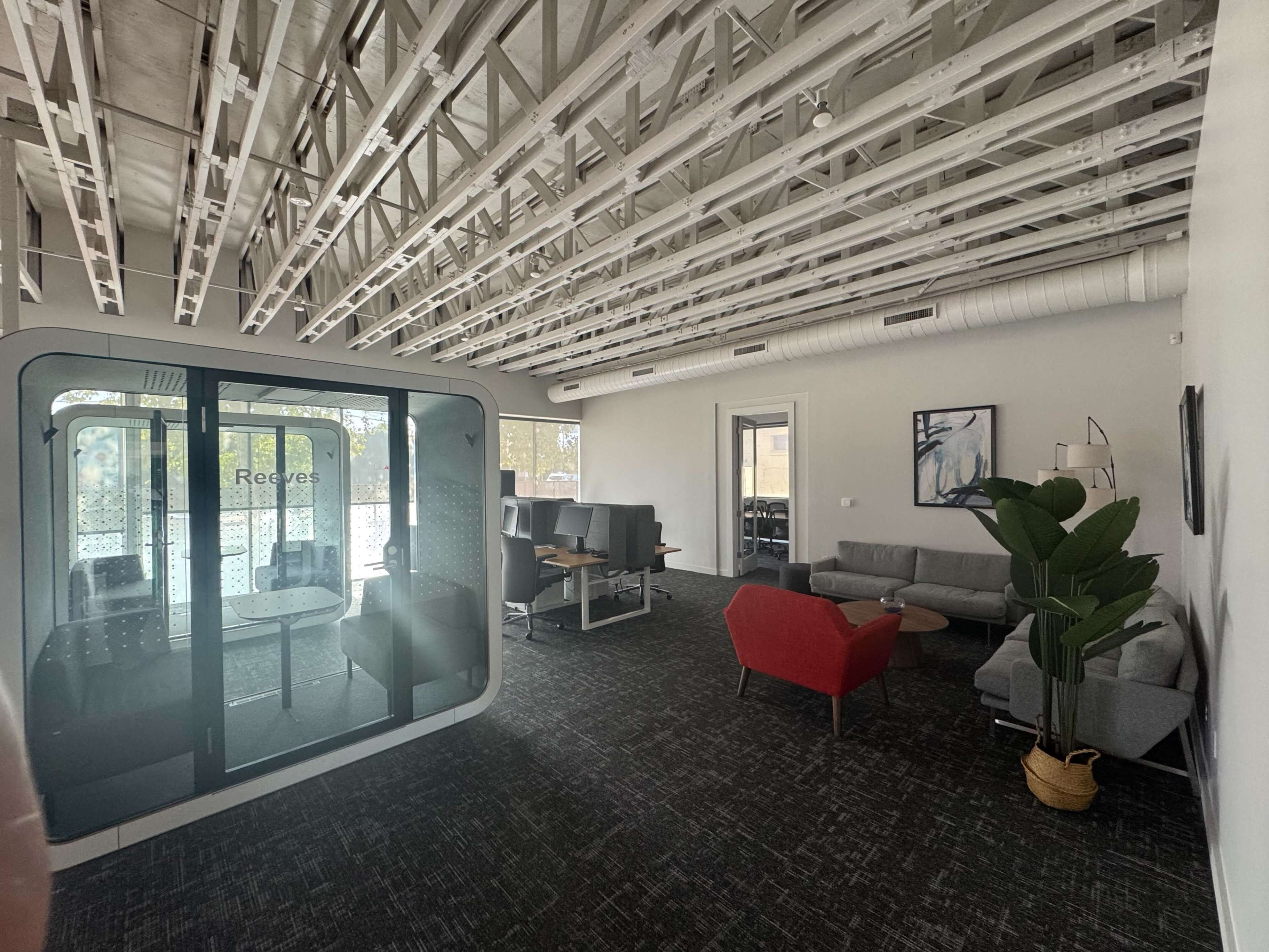 The image shows a modern office space with a glass meeting pod, seating areas, and a cable-tray ceiling.