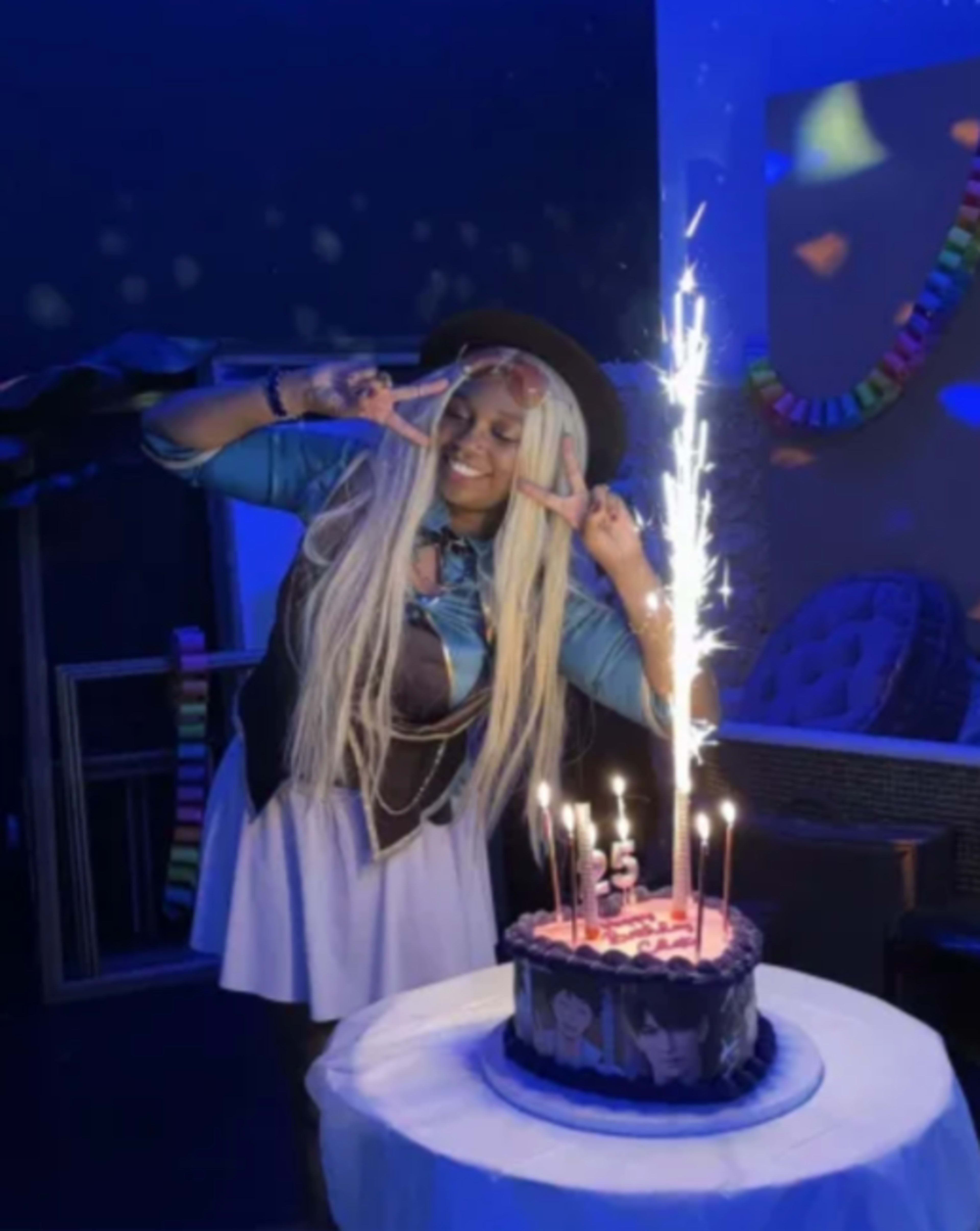A person with long blonde hair celebrates with a birthday cake featuring lit candles and sparkler fireworks in a dimly lit room.