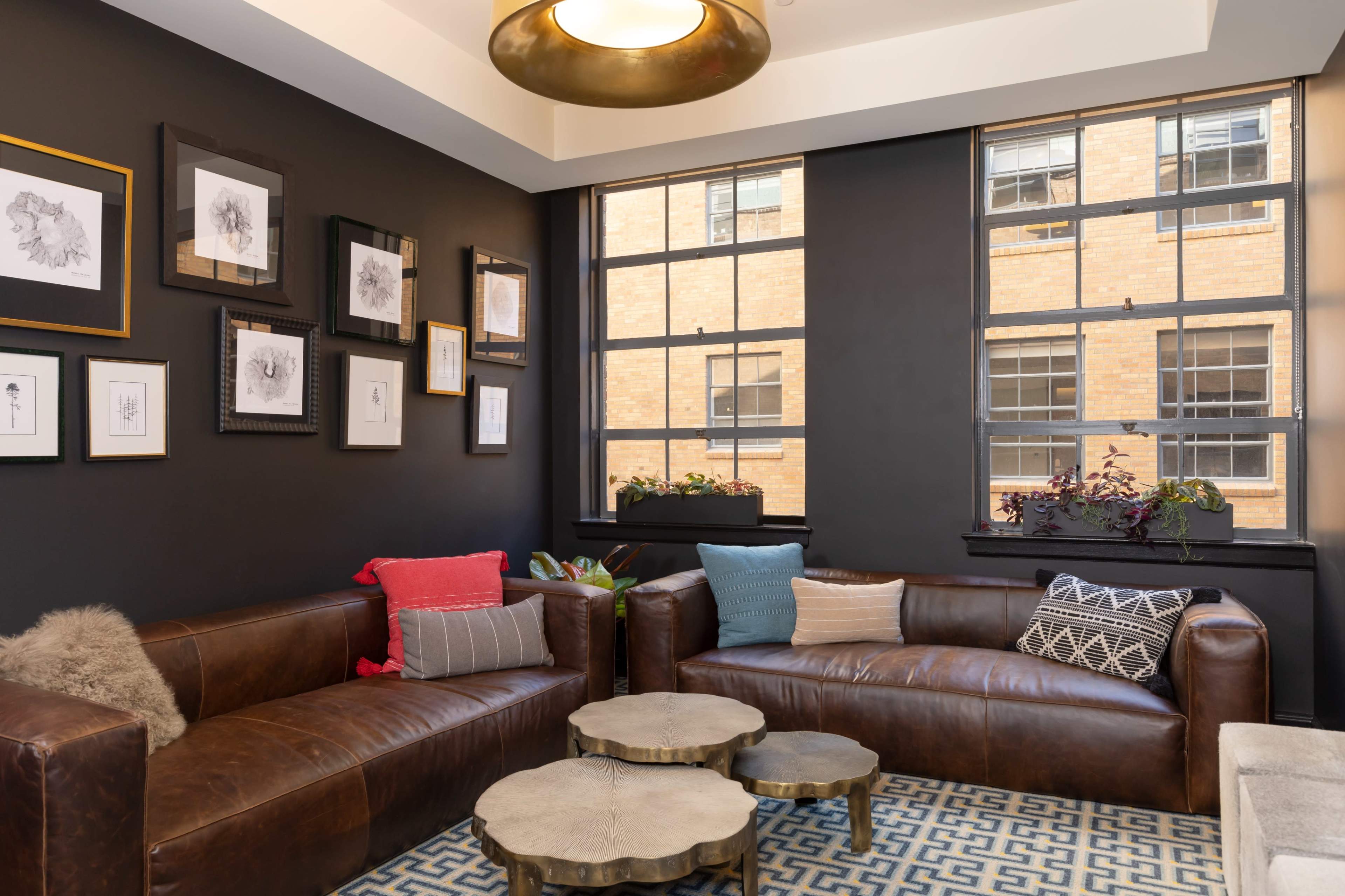 A cozy seating area features two leather sofas and a set of metallic coffee tables, illuminated by natural light streaming through large windows adorned with potted plants.