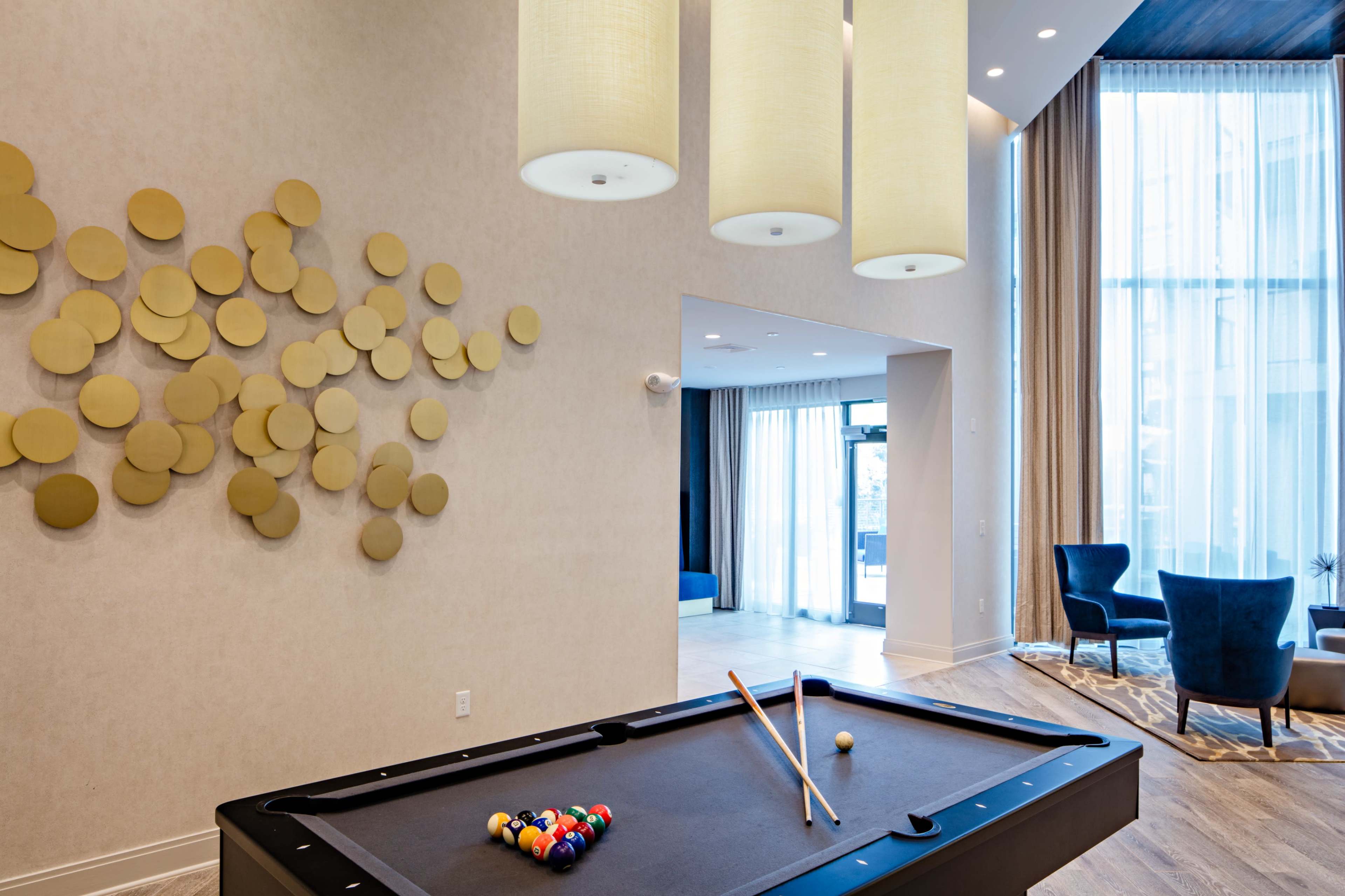 A pool table with cues and balls is set in a modern lounge featuring circular wall art and large windows draped with curtains.