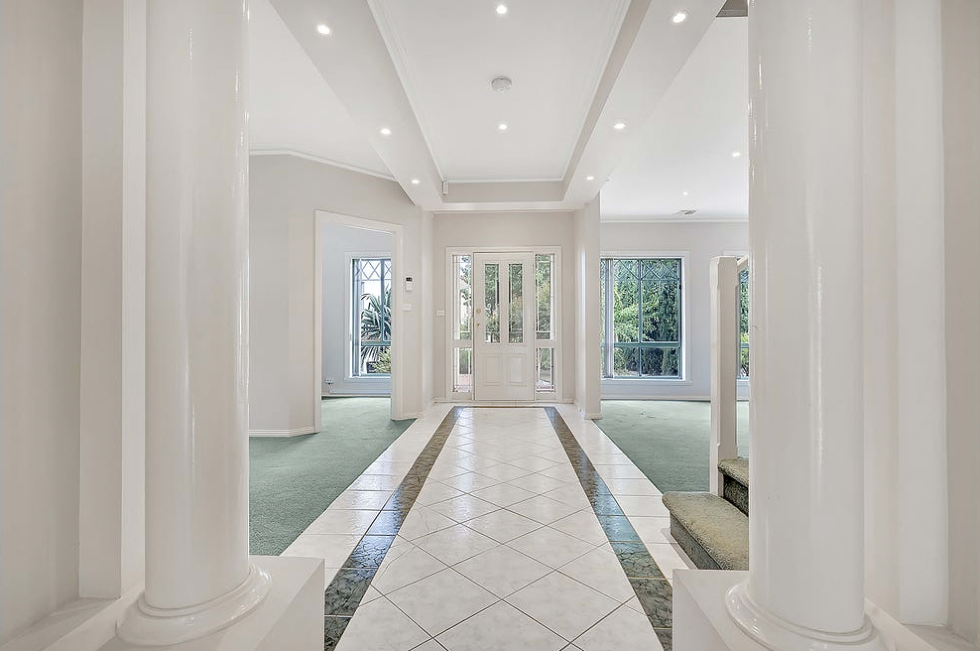 A spacious foyer features marble tiles and white columns, leading to a set of doors flanked by windows.