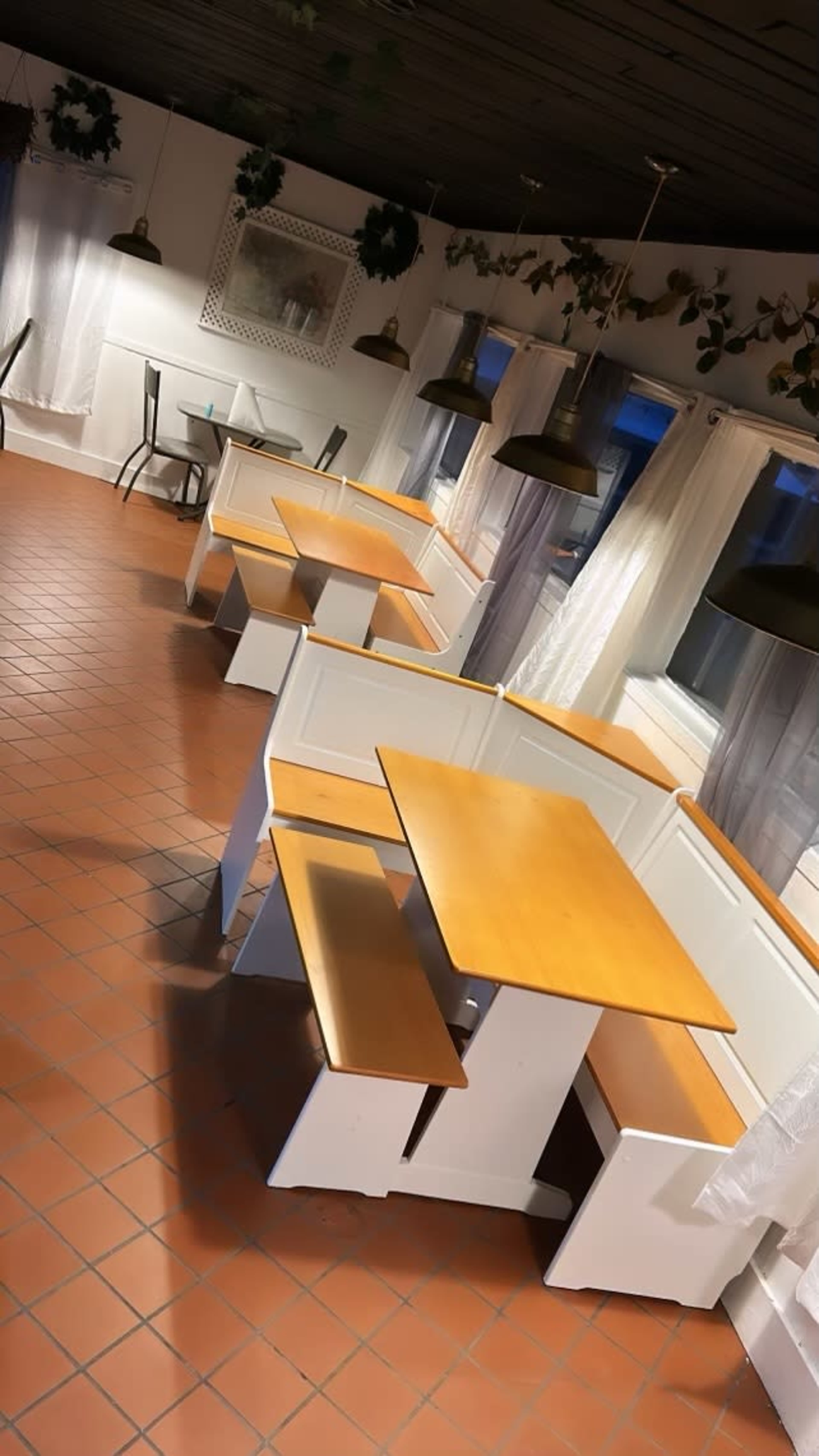 The image shows a light-filled dining area featuring multiple wooden tables and benches arranged in a square configuration, with large windows and hanging pendant lights.